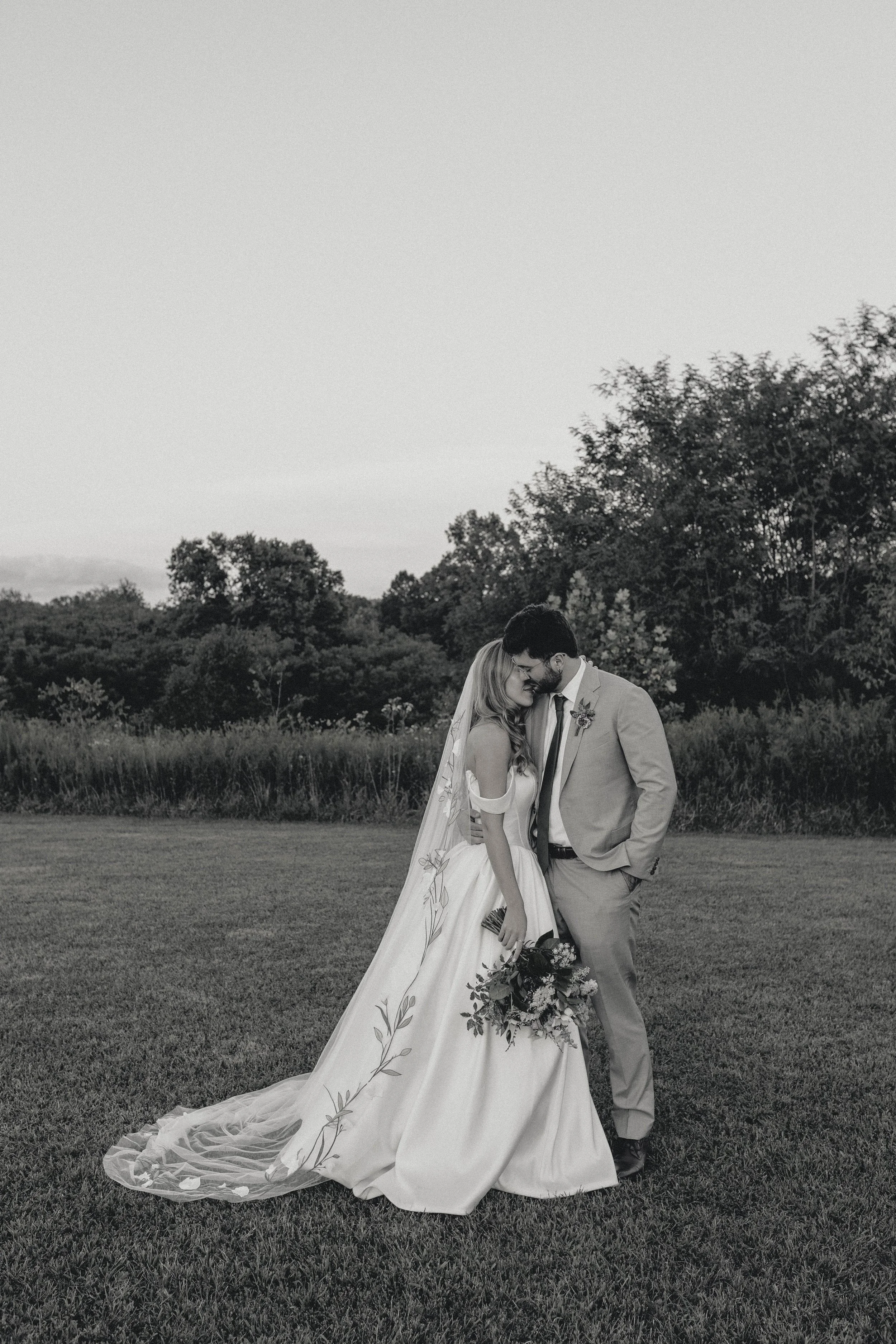 A black-and-white photo of a bride and groom standing on a grassy field, sharing a kiss. shaker village harrodsburg kentucky wedding florals