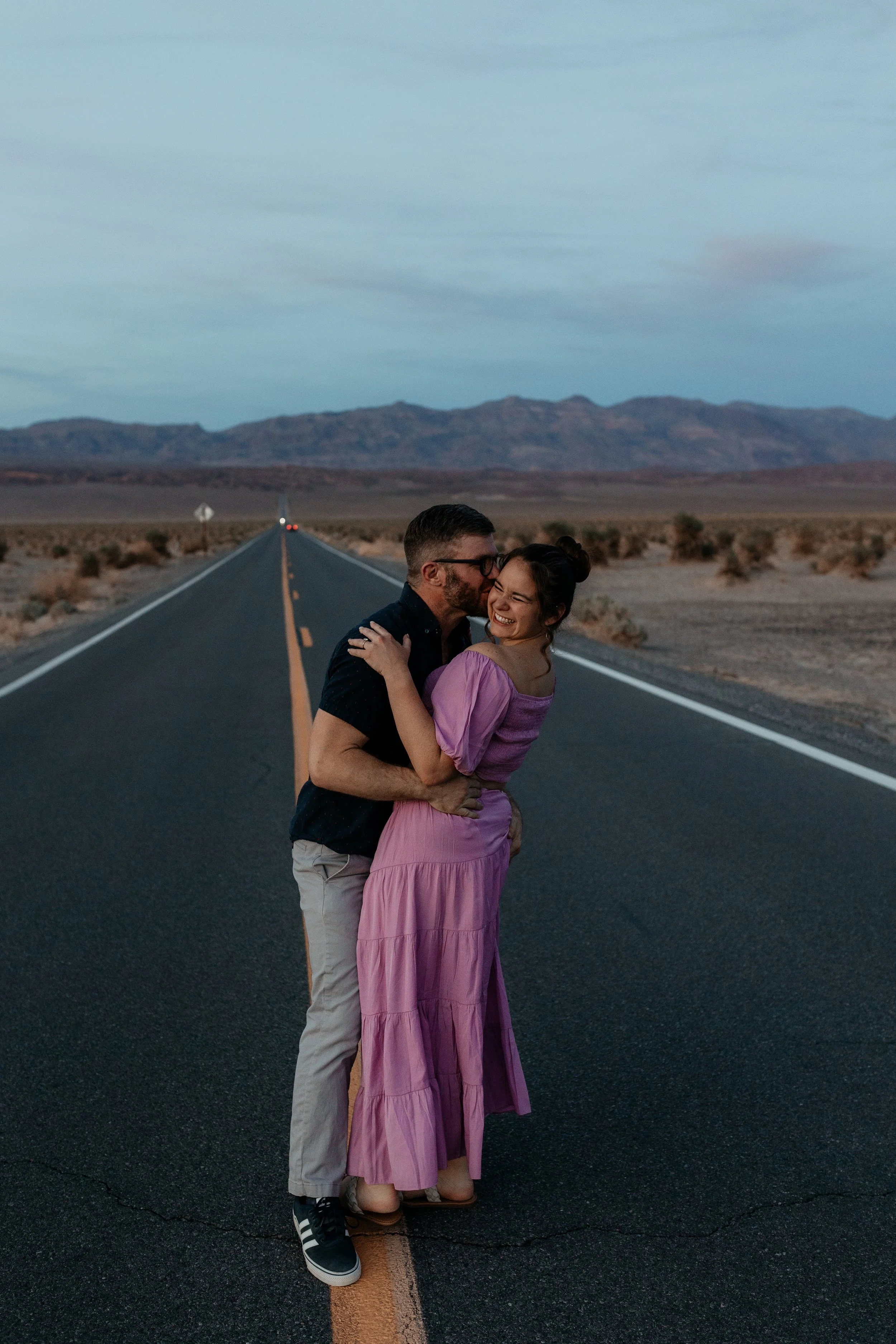 A couple hugging and kissing in the middle of an empty desert road with mountains in the background during dusk at death valley national park elopement
