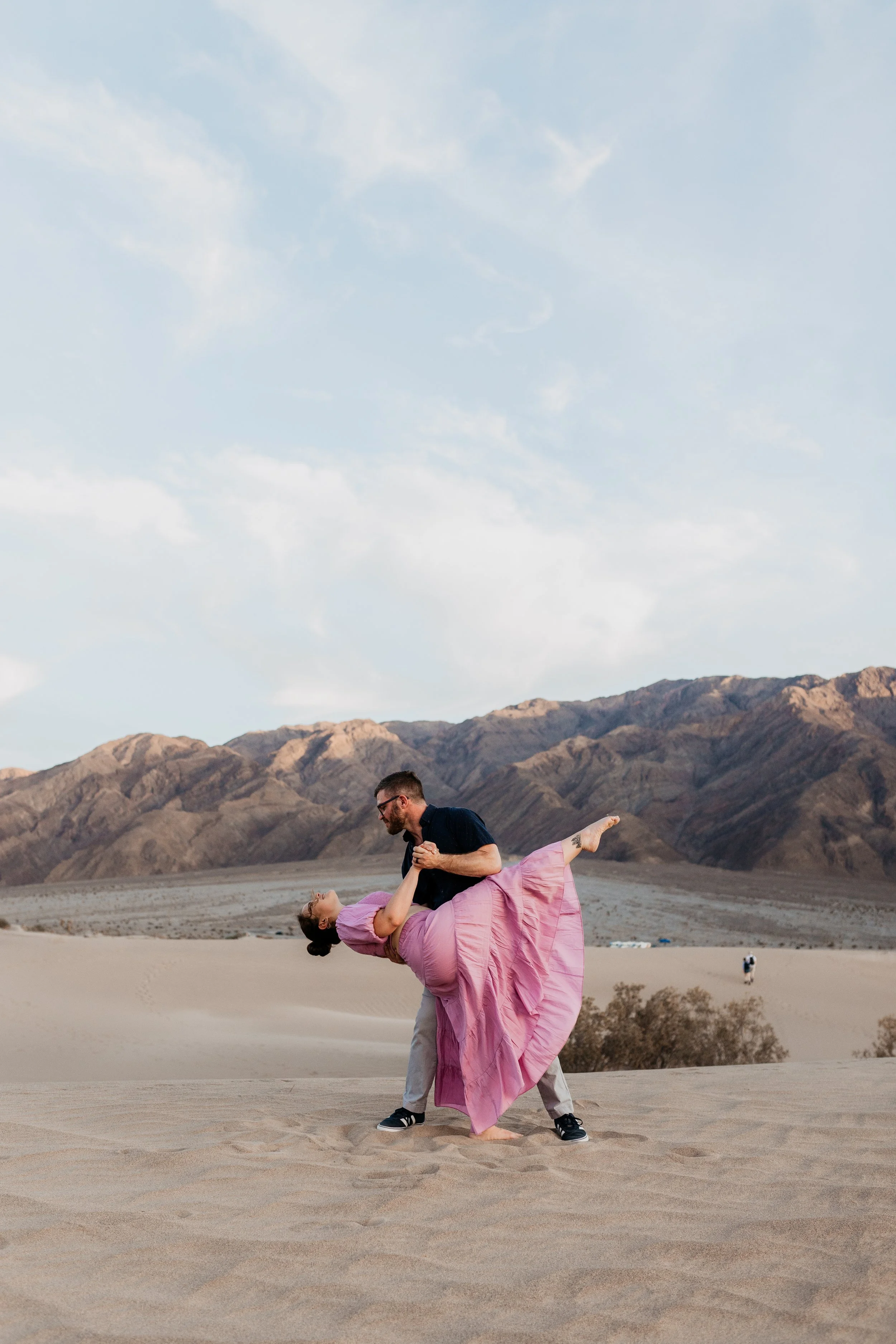 A man and woman dancing in a desert landscape with mountains and a blue sky in the background in death valley national park sand dunes