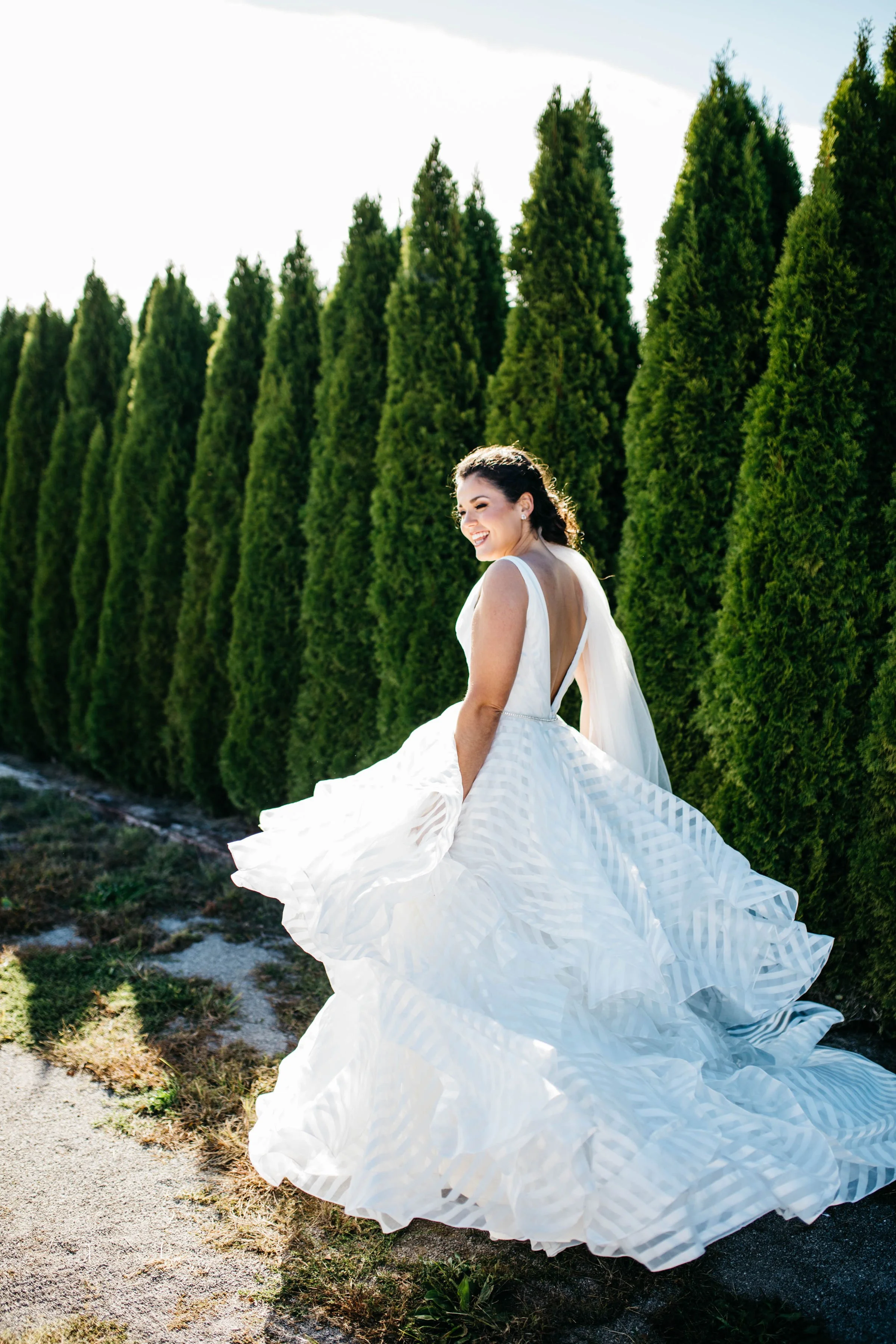 A woman in a white wedding dress outdoors, smiling, with tall green trees in the background. lexington kentucky wedding