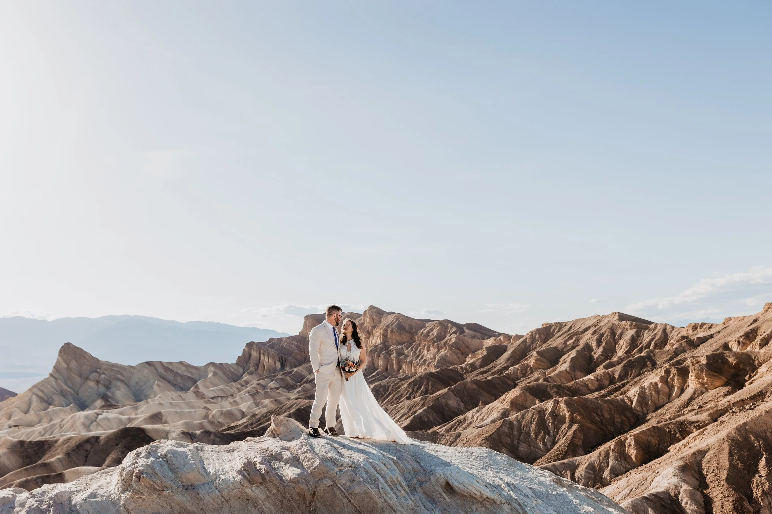 Bride and groom standing on a rock formation in a desert landscape with rugged hills under a clear sky at death valley national park elopement