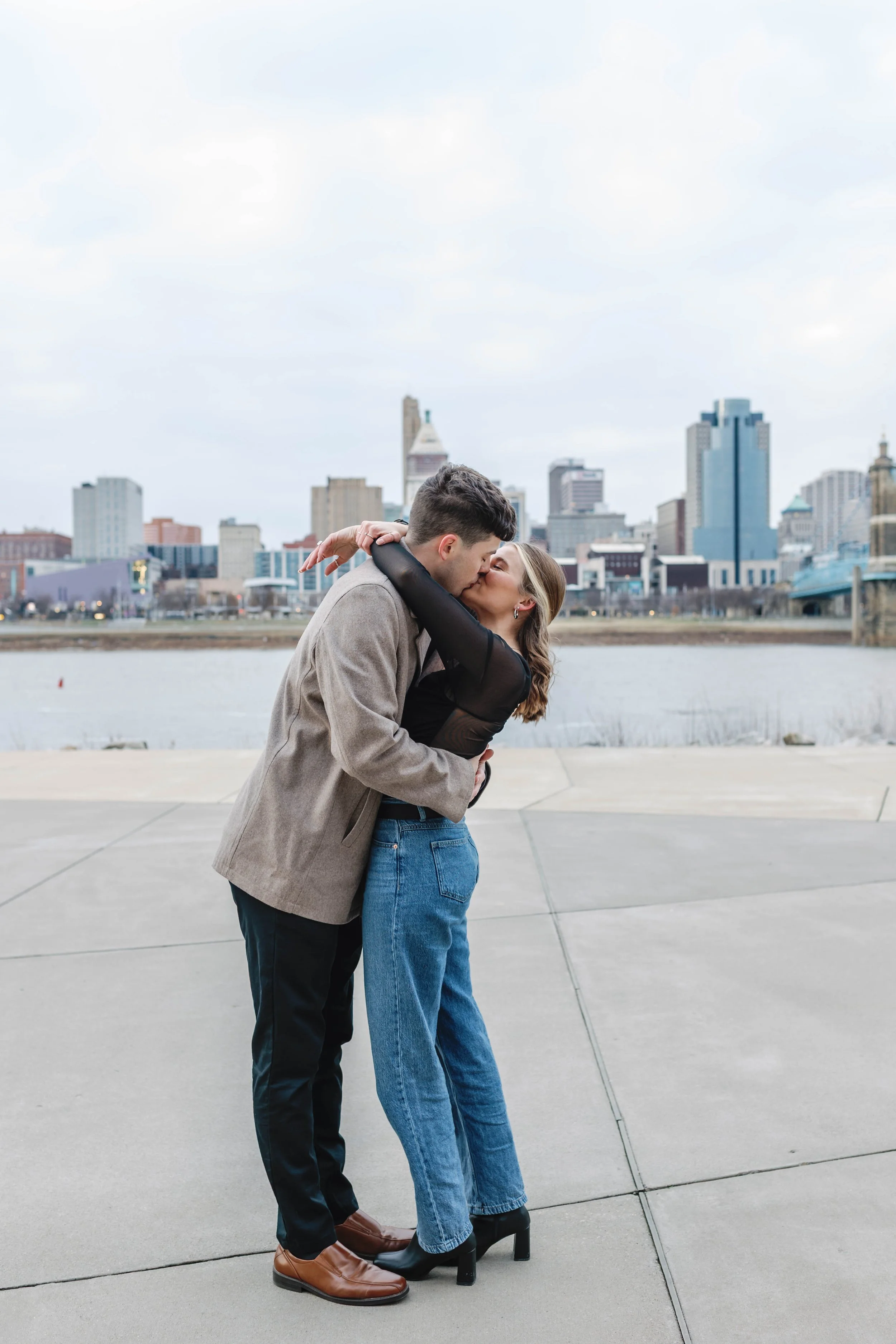 A couple kissing by a river with city skyline in the background. proposal at covington riverwalk with cincinnati skyline in background engagement 