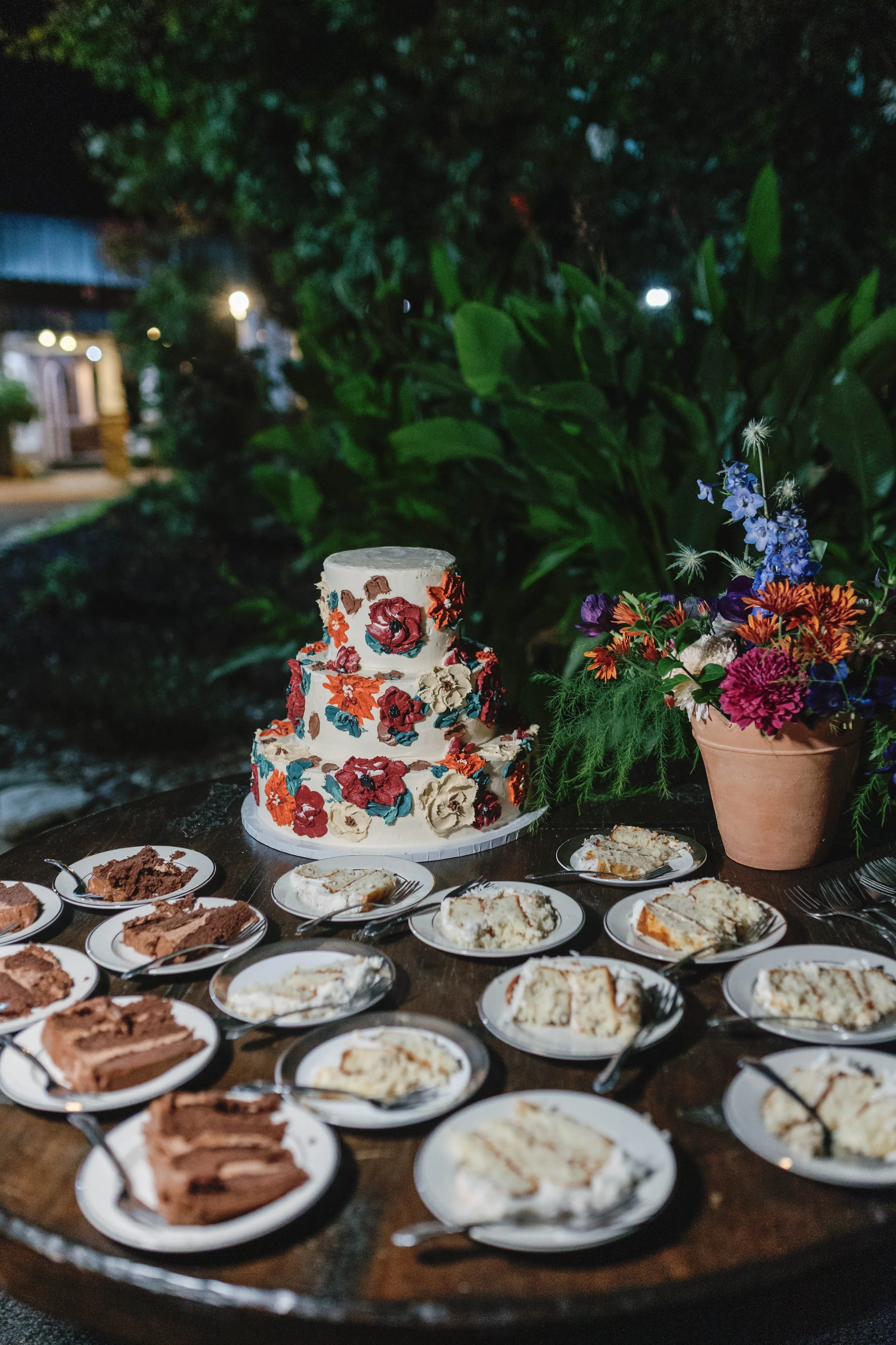 A tiered floral decorated cake on a table surrounded by slices of cake on plates, with a large vase of colorful flowers beside it, outdoors at night. elegant kentucky backyard wedding 