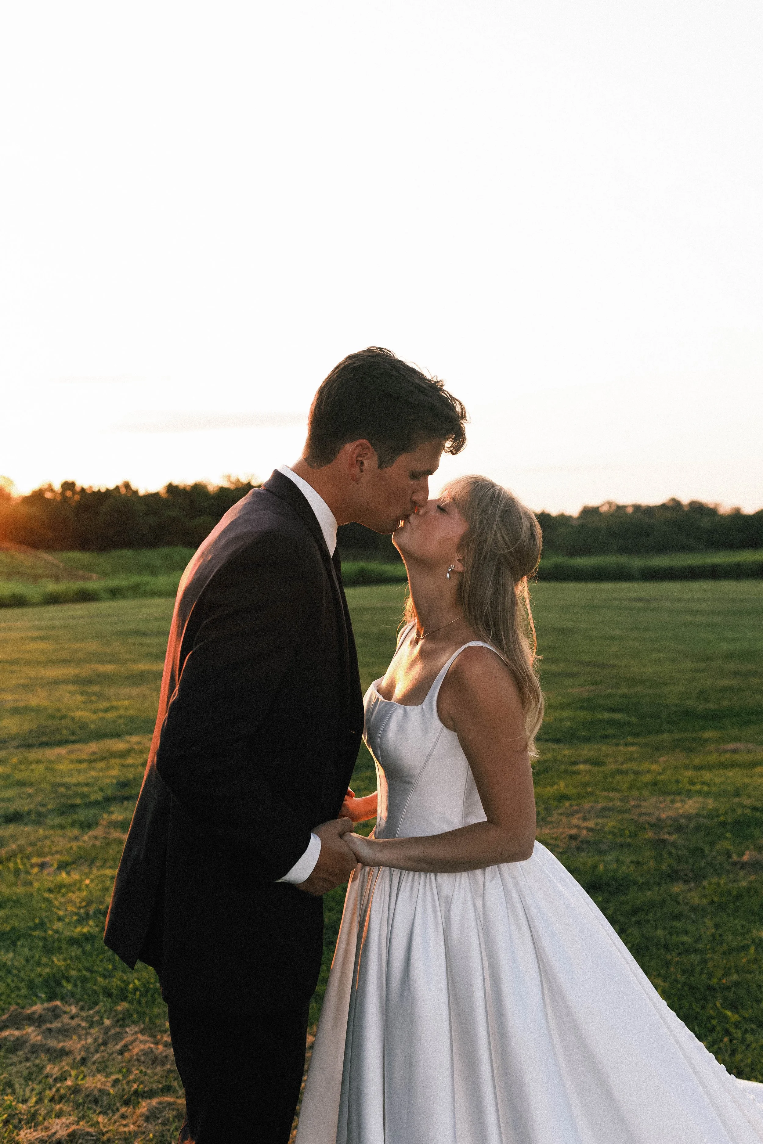 A newlywed couple sharing a kiss outdoors at sunset, holding hands, with a grassy field and trees in the background. ashford acres inn wedding cynthiana kentucky
