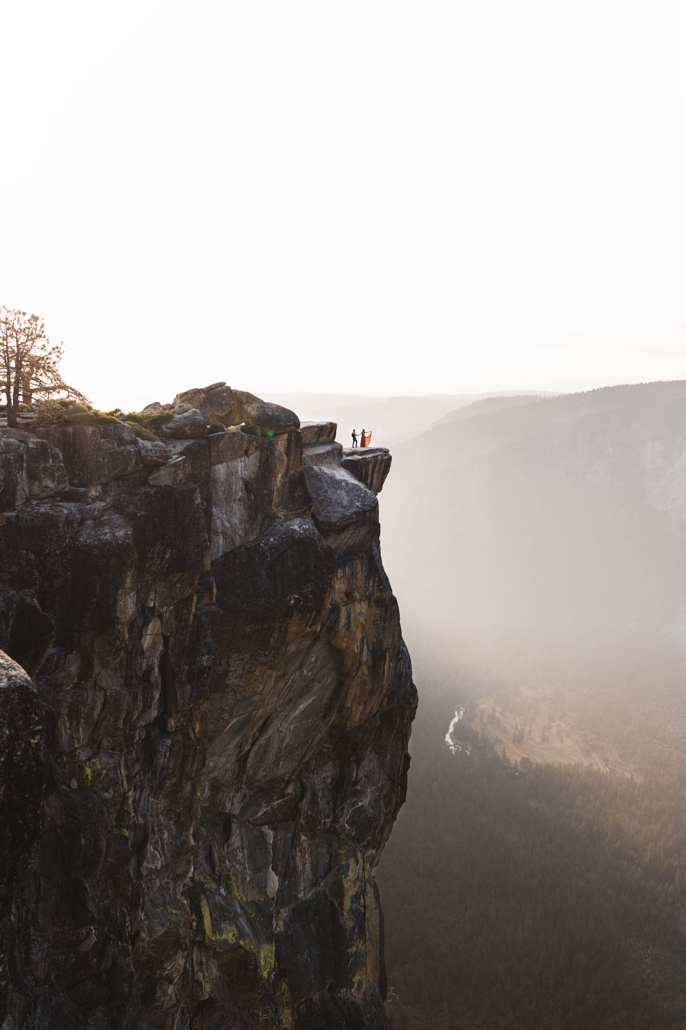 Two people standing on the edge of a high cliff, overlooking a valley with a river and forested landscape, during sunrise or sunset at taft point in yosemite national park california elopement