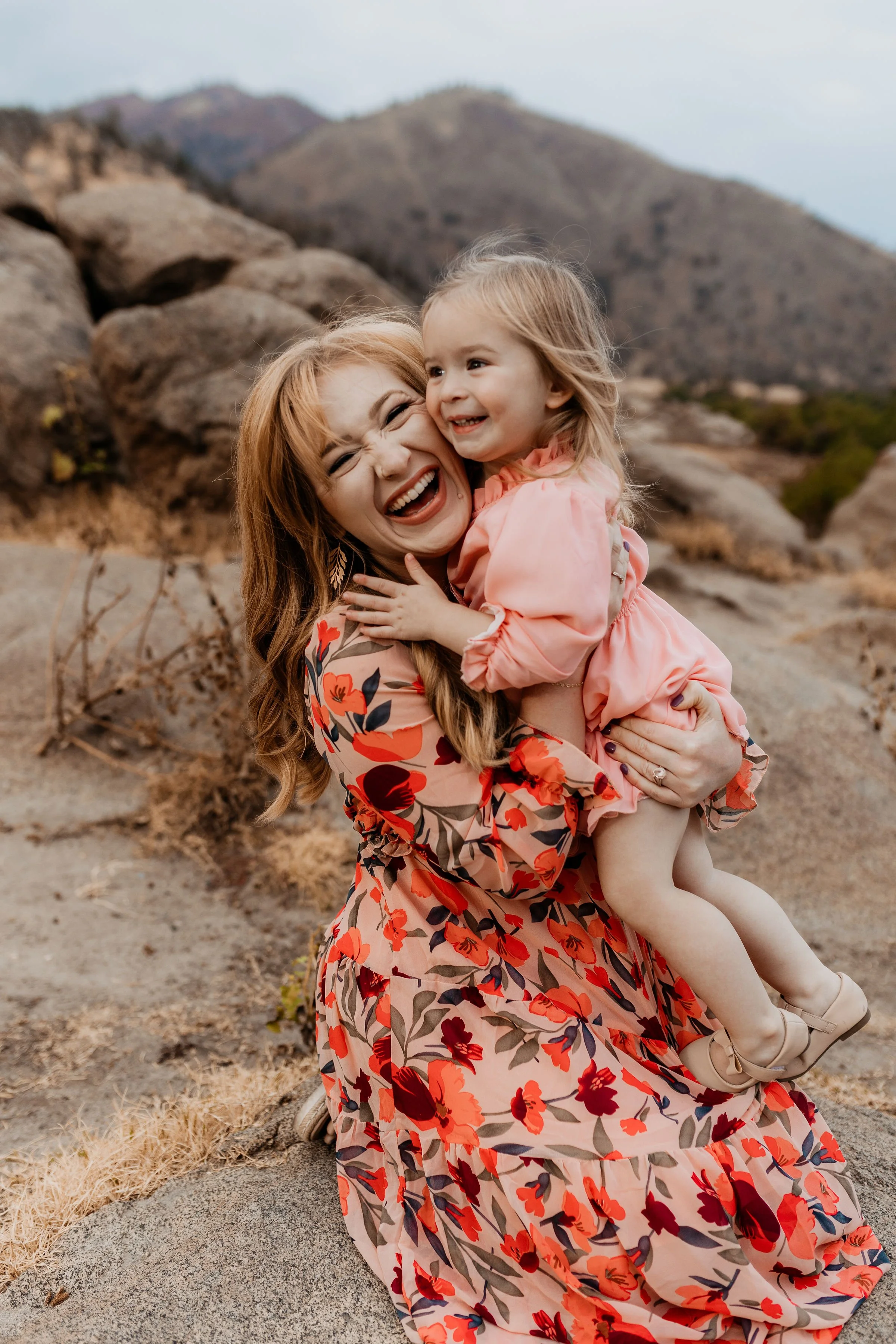 A woman with red hair in a floral dress holding a young girl in a pink dress outdoors in a desert landscape with mountains in the background, both smiling and laughing.  lexington kentucky family photographer storytelling portraits
