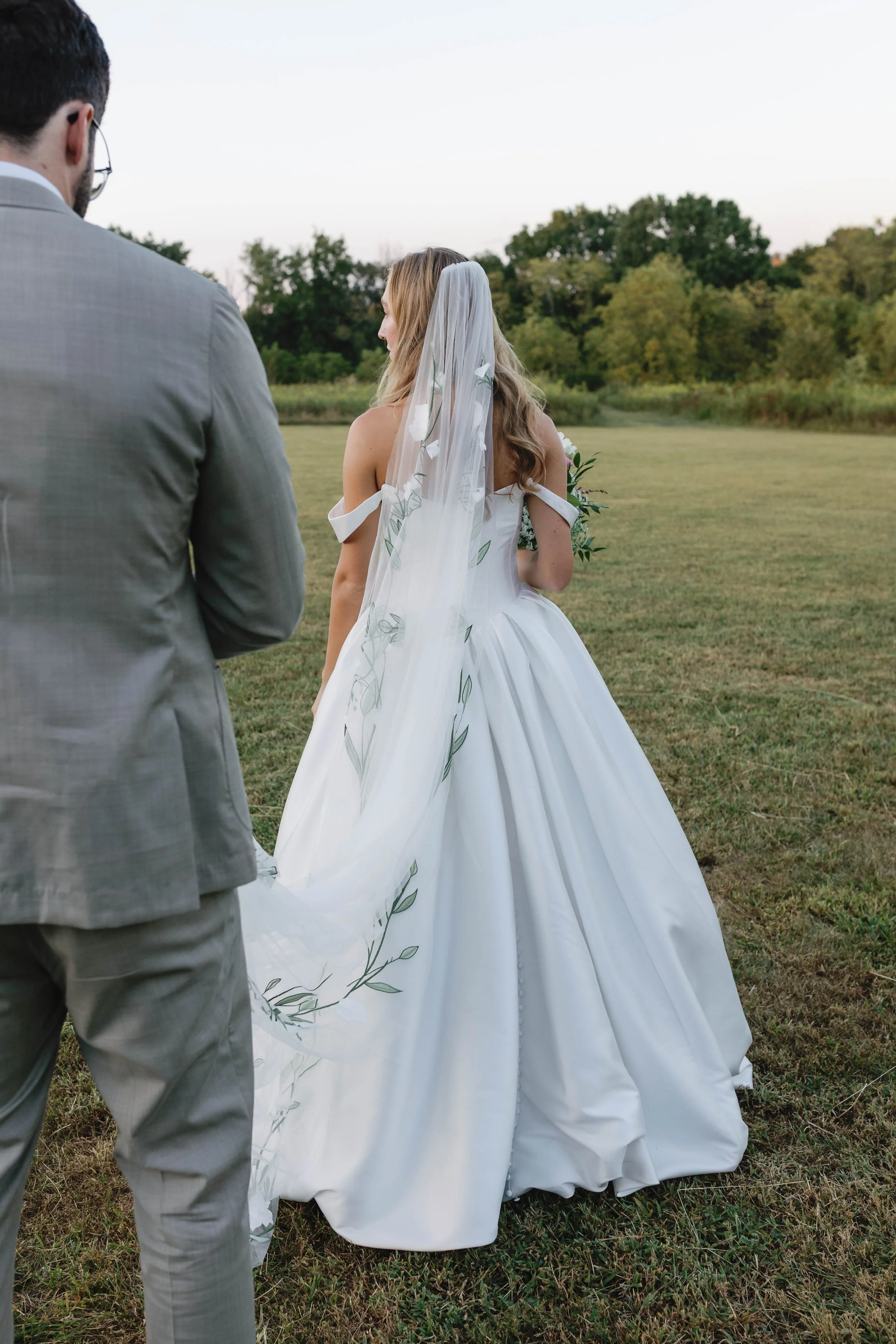 A bride in a white wedding gown and veil stands outside on a grassy field, with a man in a gray suit facing her during sunset or late afternoon. shaker village of pleasant hill kentucky wedding summer