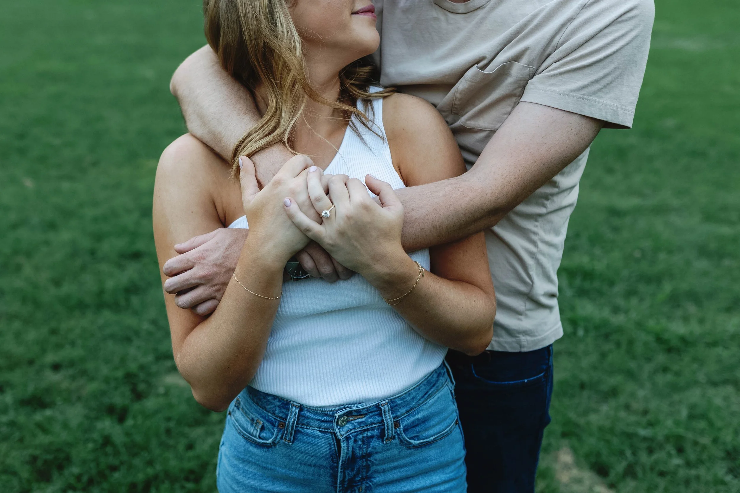 A couple embraces outdoors on green grass; the woman wears a white sleeveless top and denim jeans, and the man wears a beige T-shirt engagement photos at park in Louisville kentucky
