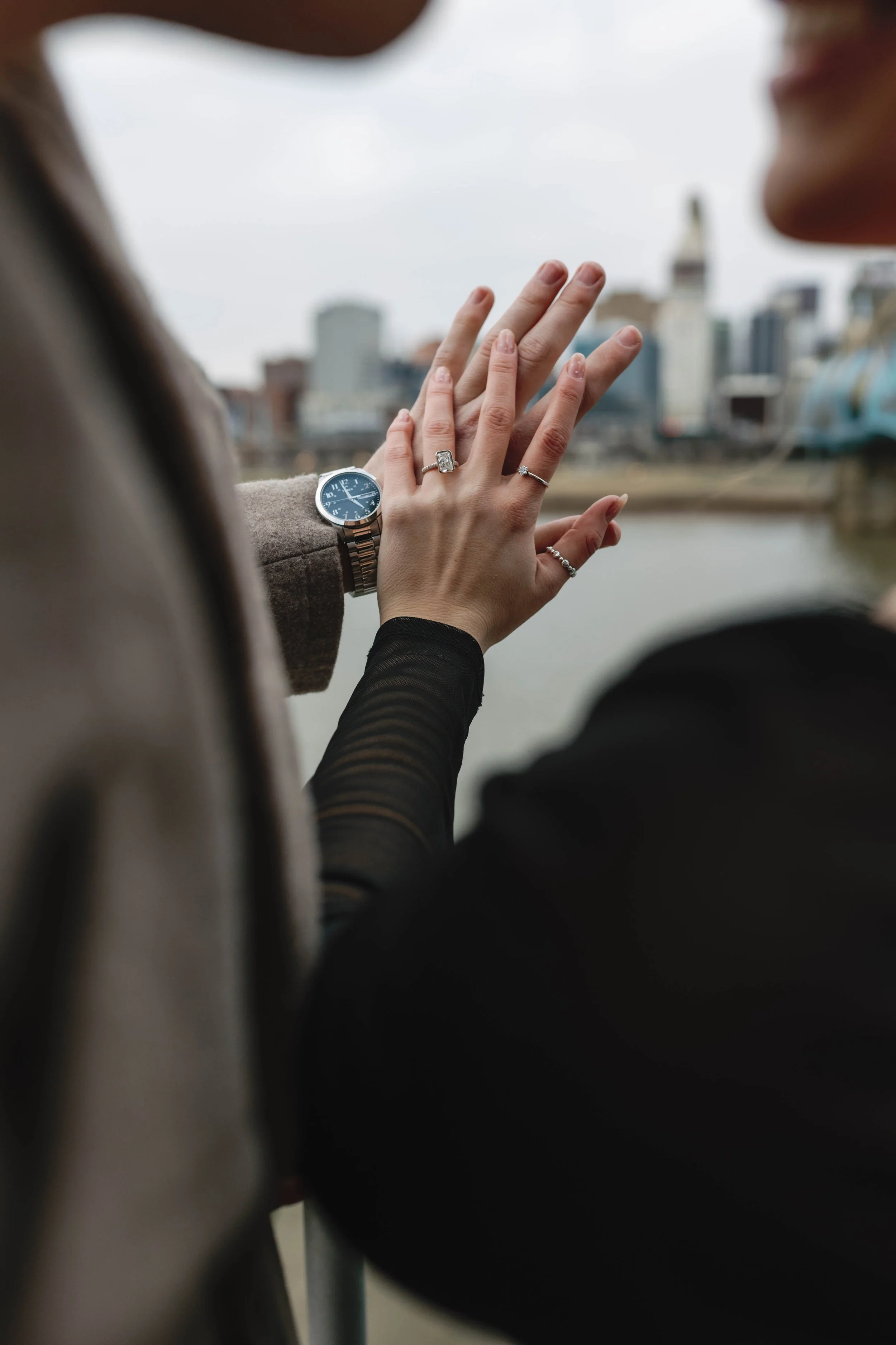 Close-up of two women holding hands with wedding rings against a cityscape and river background, one wearing a watch. proposal at covington riverwalk with cincinnati skyline in background engagement 