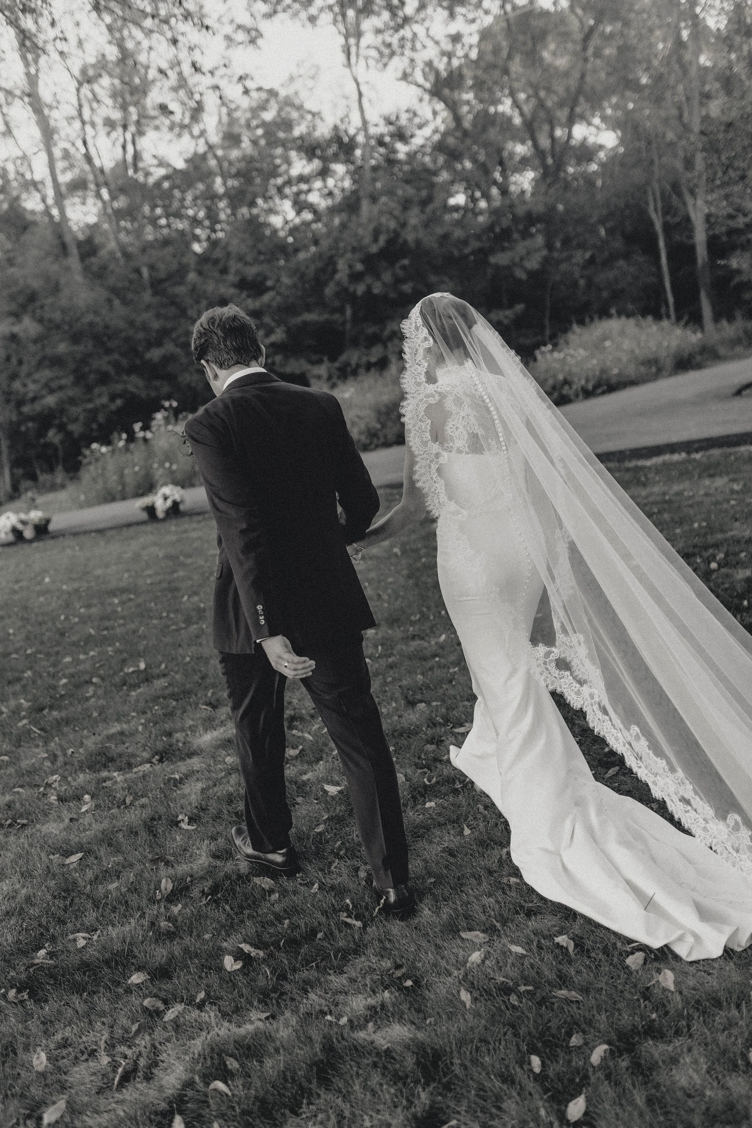A bride and groom walking outdoors on a grassy area, holding hands, with trees and flowers in the background. The bride wears a long dress with lace details and a veil, while the groom is in a black suit. kentucky elegant wedding