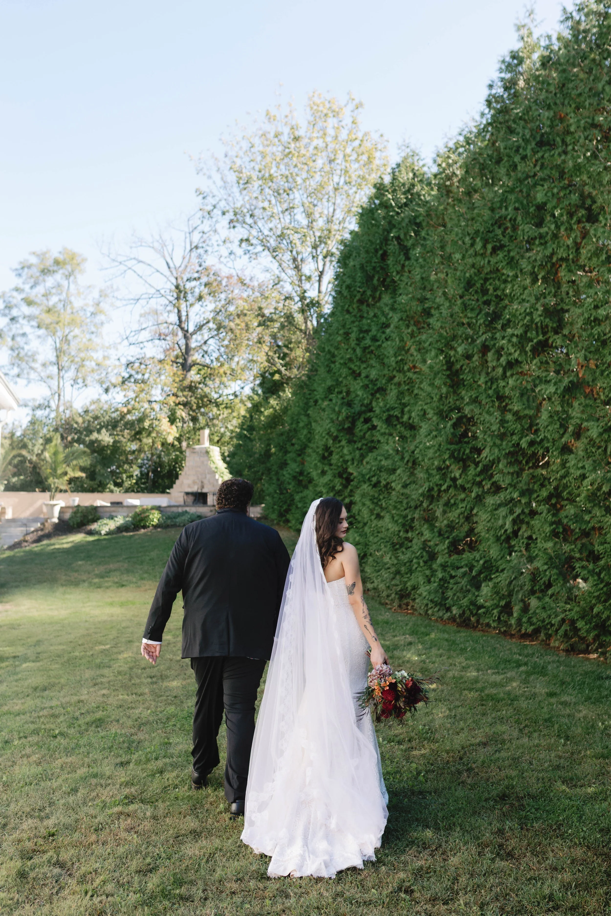 A bride in a wedding dress with a veil holding a bouquet of flowers walking with a groom dressed in a black suit outdoors on a grassy lawn with tall green bushes and trees in the background. moundale manor historic kentucky wedding venue winchester k
