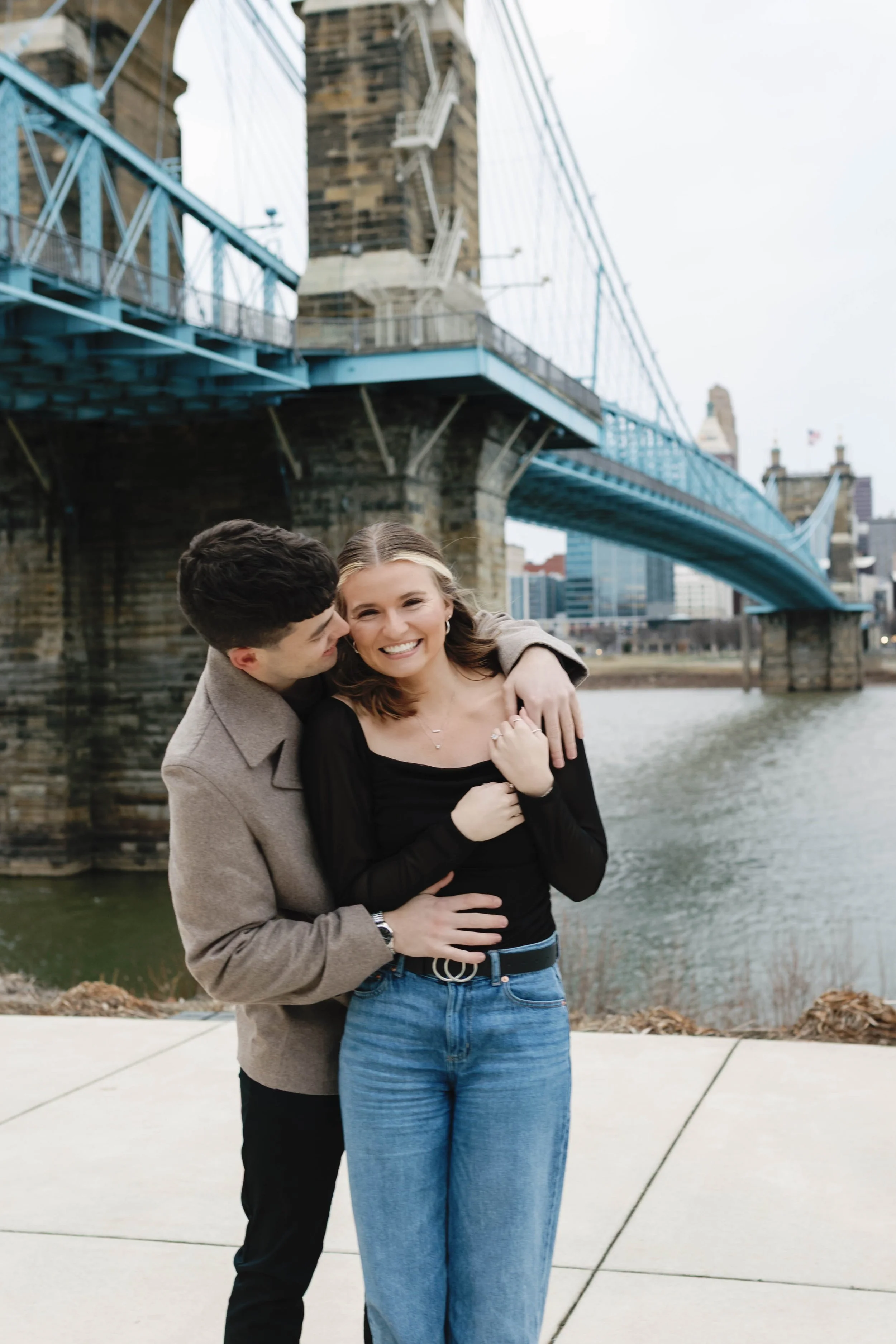 A young couple sharing a hug and smiling near a river with a blue suspension bridge in the background proposal at covington riverwalk with cincinnati skyline in background engagement 