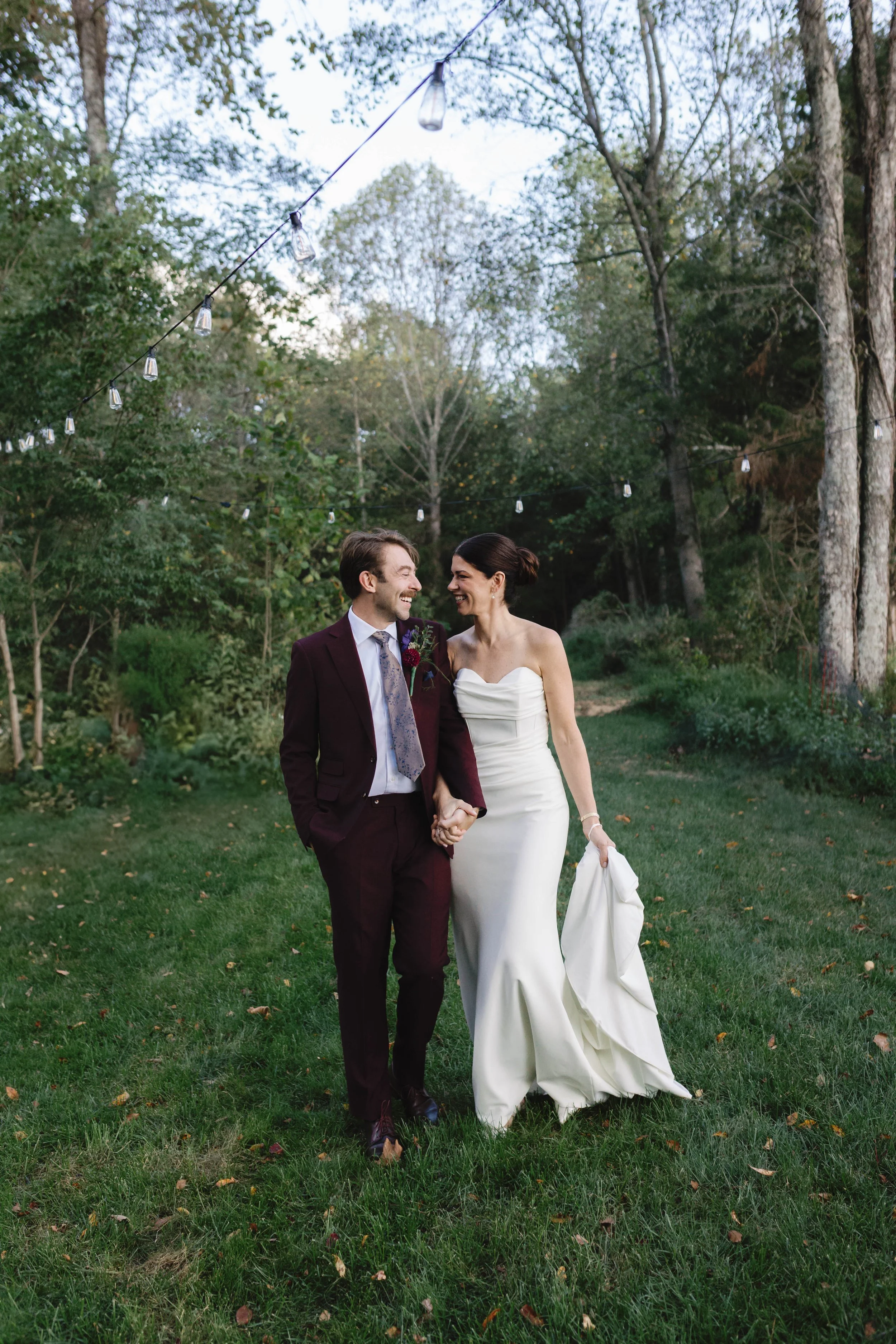 A newlywed couple walking hand in hand outdoors at dusk, surrounded by trees and string lights overhead, in a joyful moment elegant backyard kentucky wedding