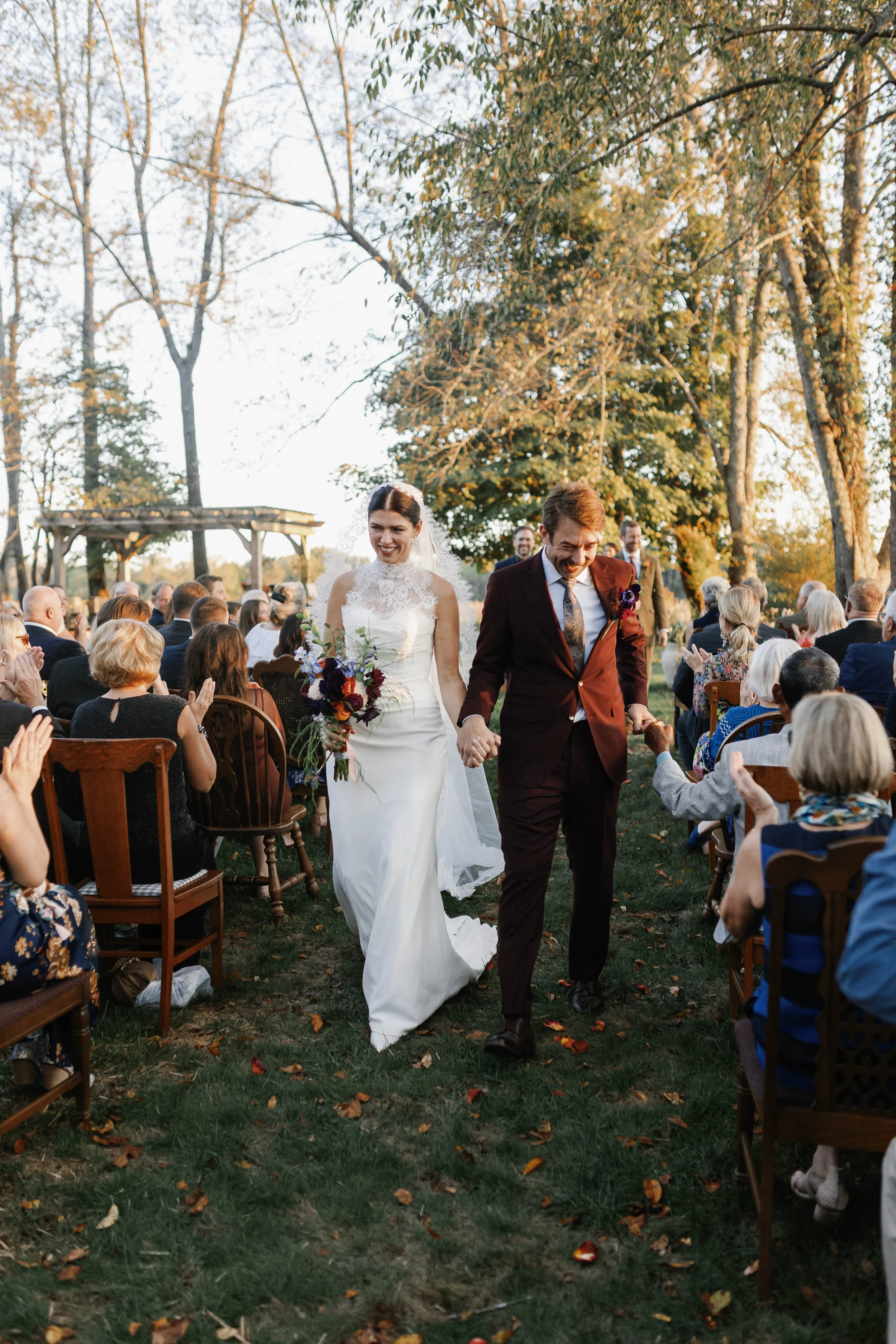 Bride and groom walking hand-in-hand down the aisle at an outdoor wedding ceremony, surrounded by seated guests in a wooded area with autumn leaves. elegant kentucky backyard wedding 