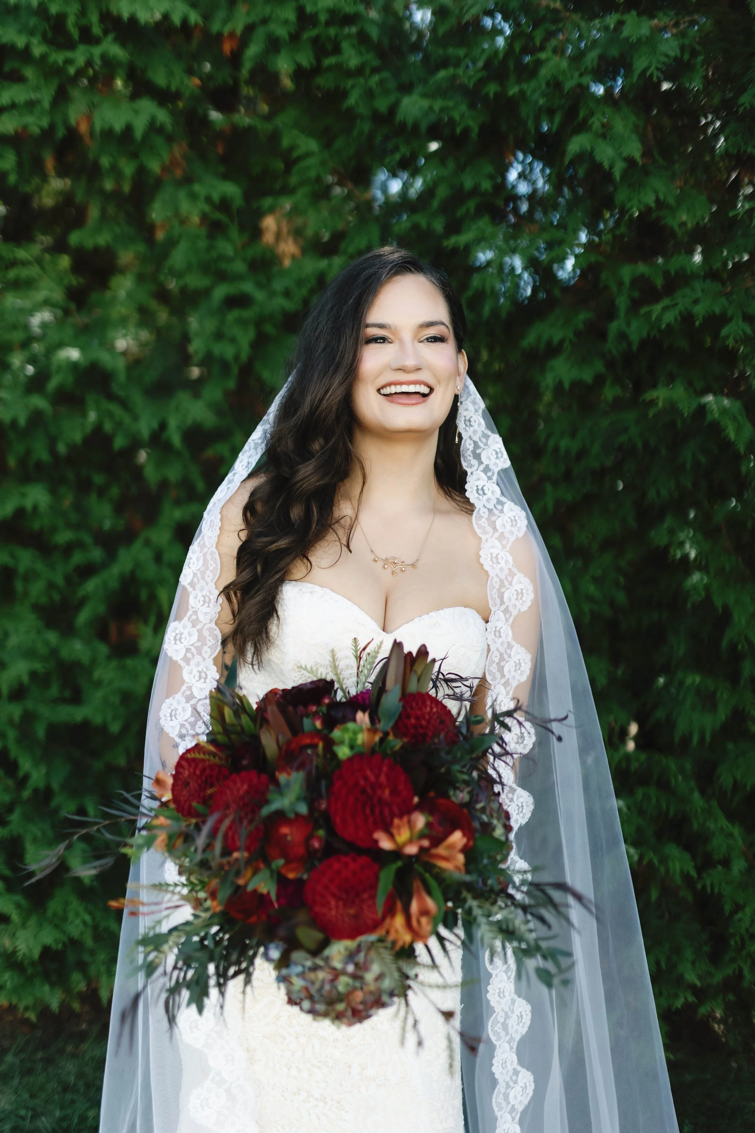 Bride holding a large bouquet of red flowers with greenery, smiling, standing in front of green trees in Moundale Manor historic wedding venue winchester kentucky