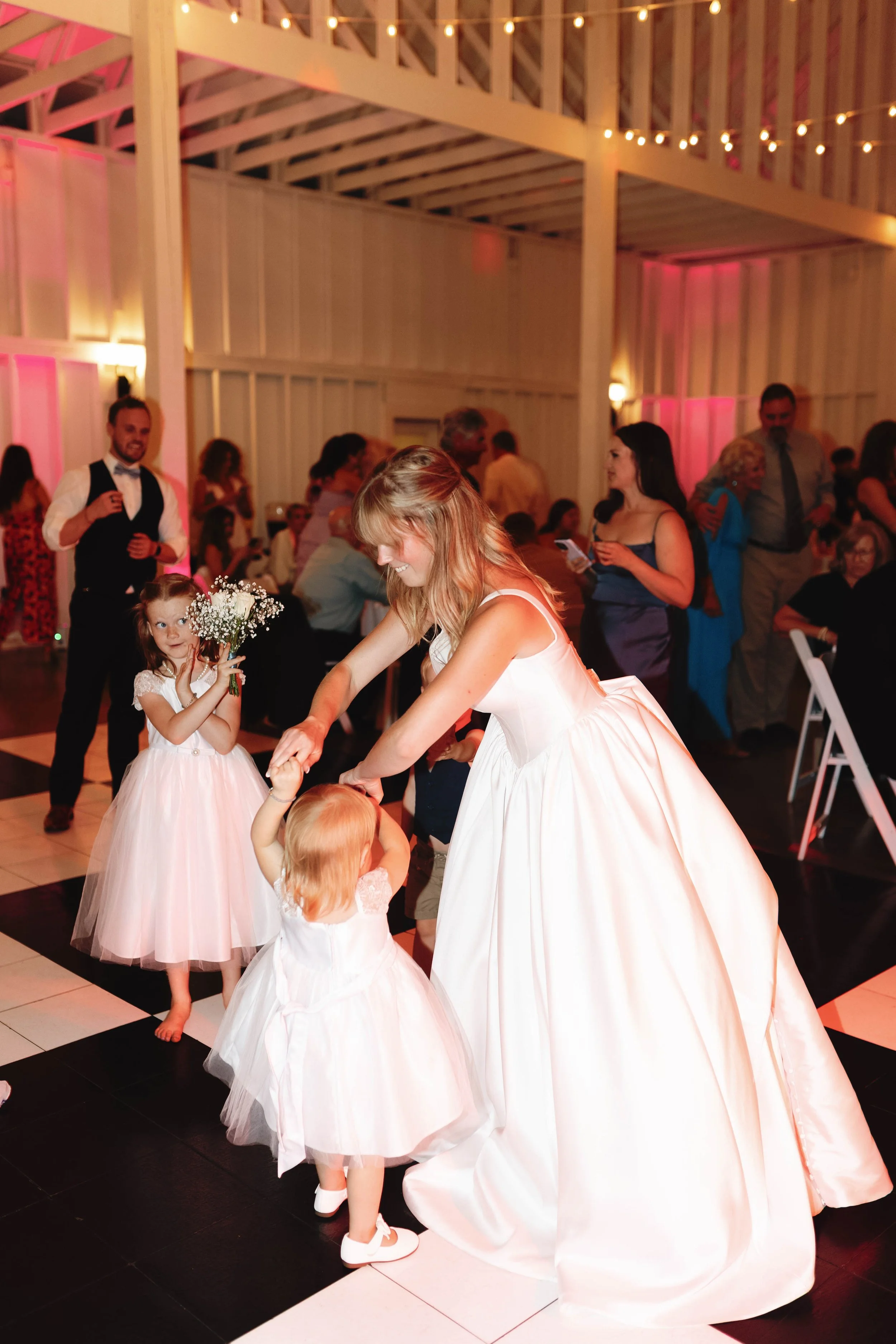A woman in a white wedding dress dancing with two young girls in white dresses at a wedding reception, with other guests in the background. ashford acres inn wedding cynthiana kentucky