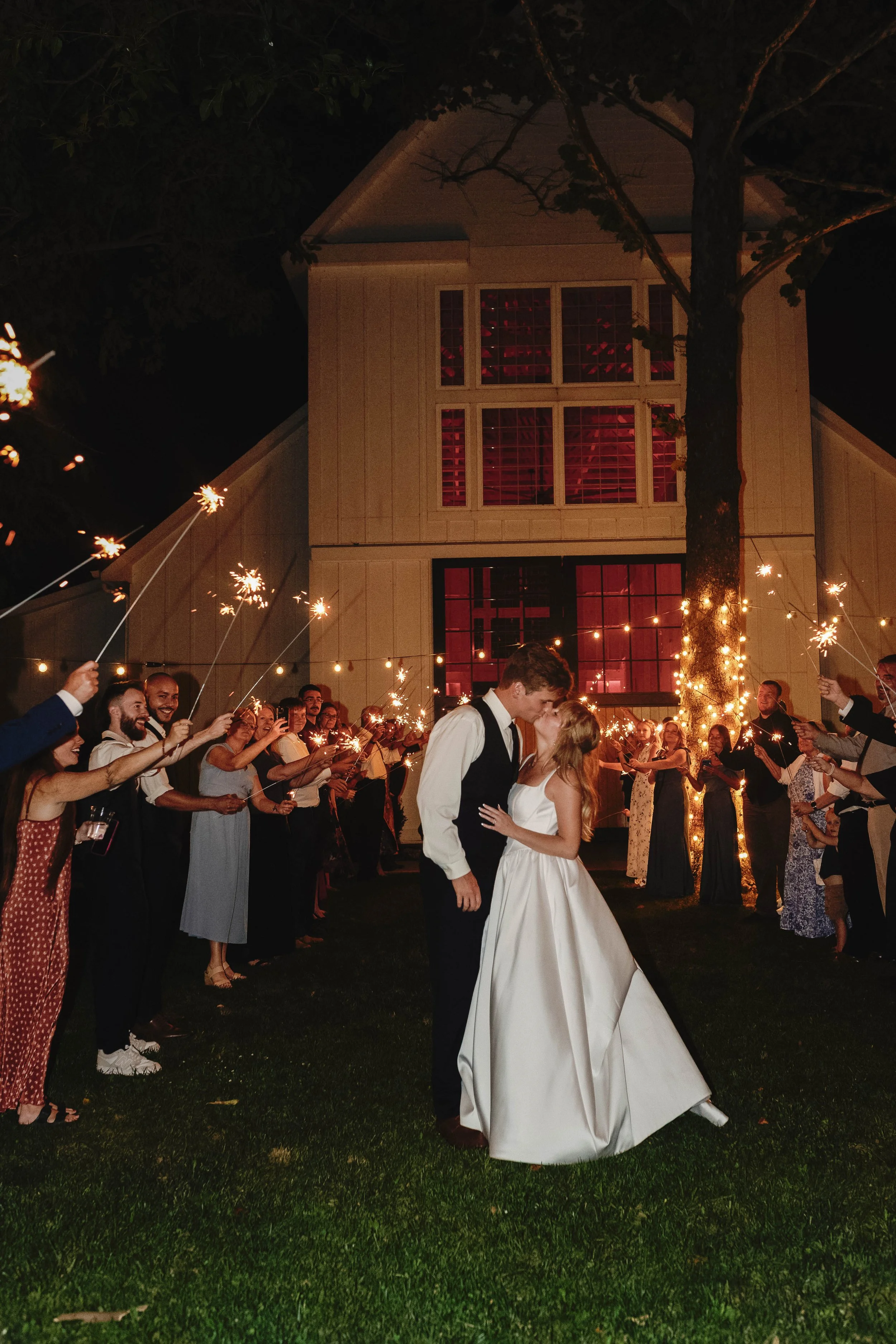 Couple in wedding attire kissing under string lights, surrounded by guests holding sparklers at night outside a house. ashford acres inn wedding cynthiana kentucky
