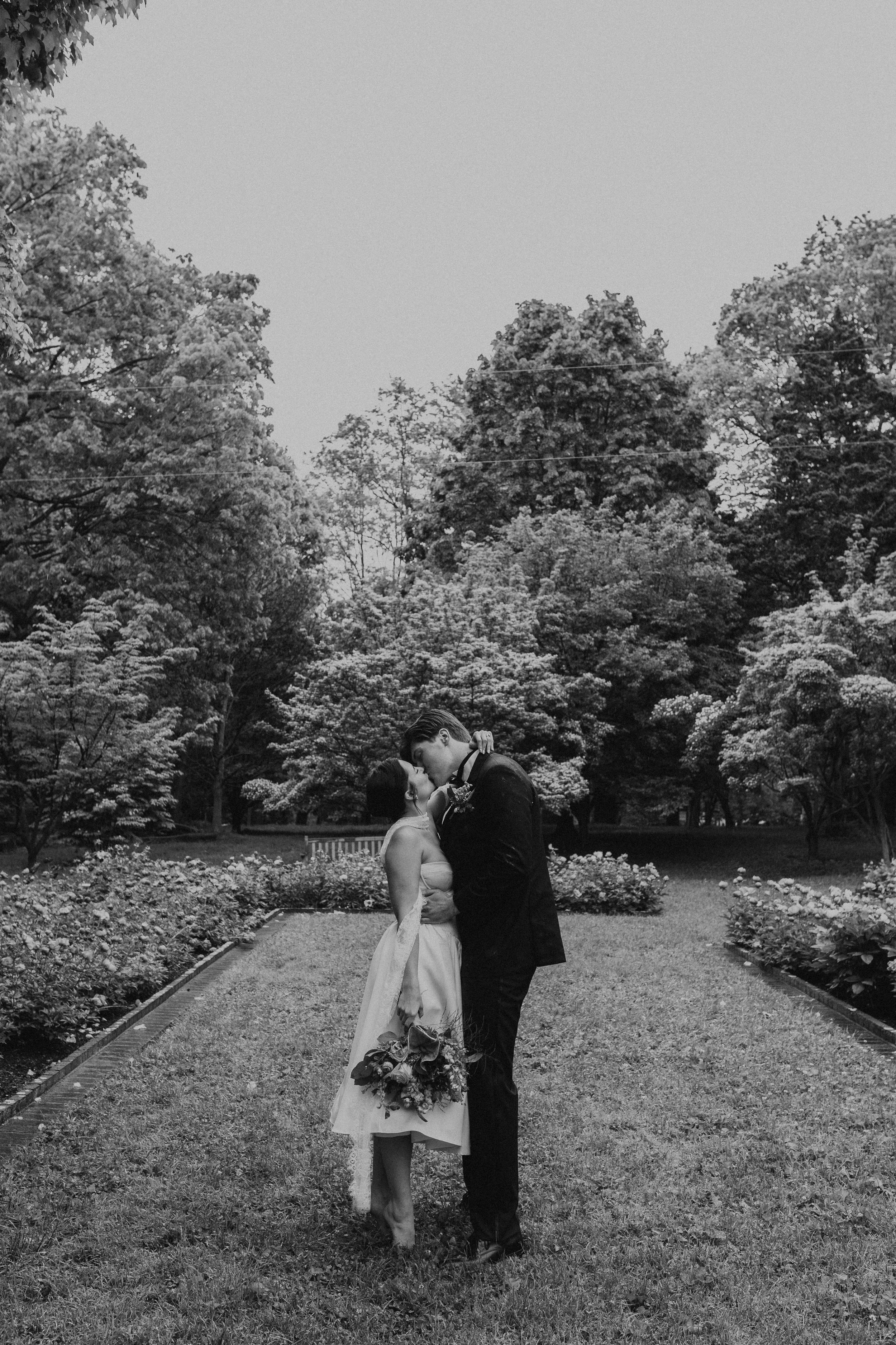 A couple in wedding attire sharing a kiss in a garden with trees and flowers. Ashland Henry Clay wedding lexington kentucky