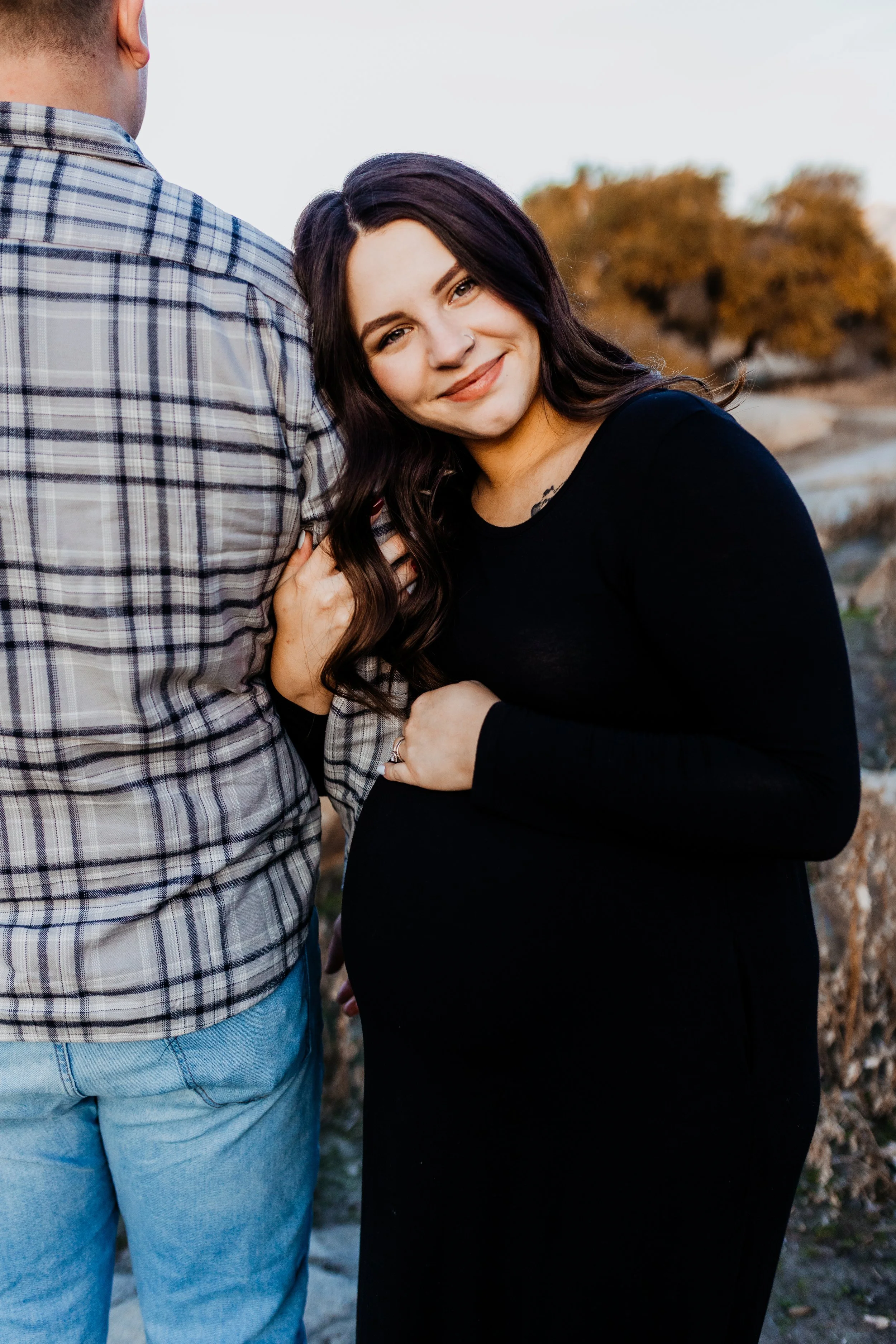 A pregnant woman with long dark hair smiling and hugging a man in an outdoor setting with trees and rocks in the background.  lexington kentucky family photographer storytelling portraits maternity 