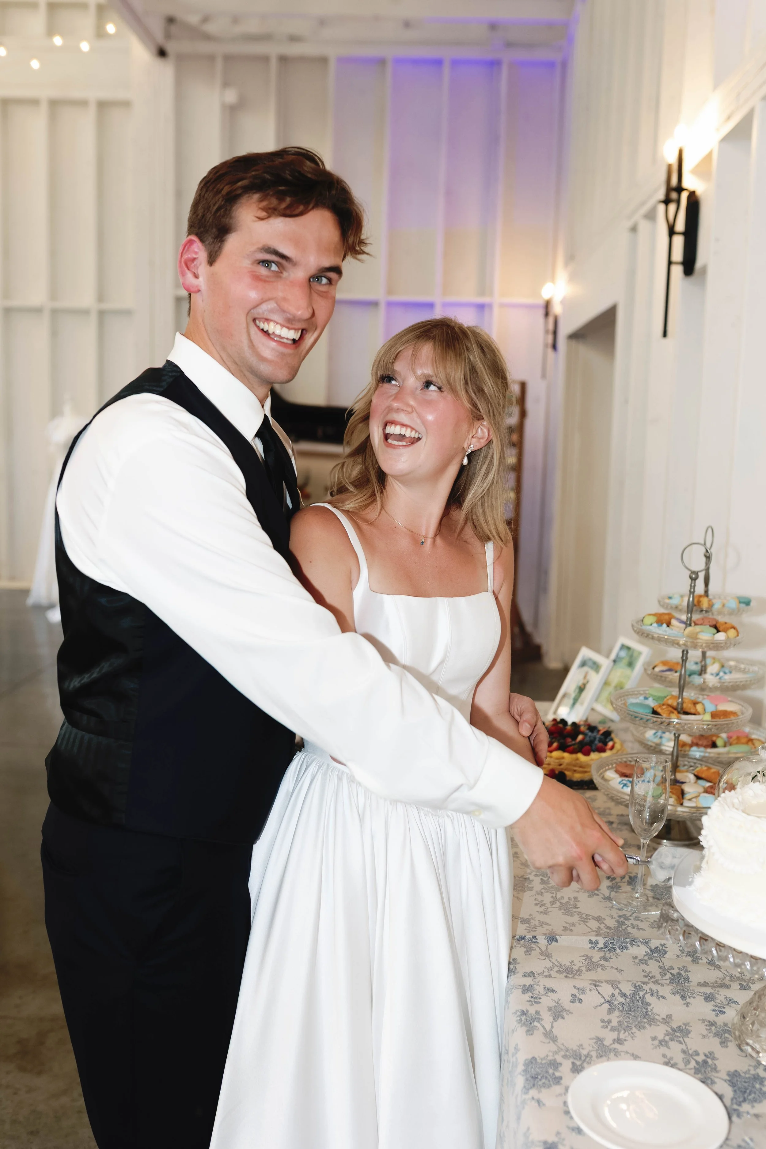 A couple at a wedding reception, cutting a wedding cake together, smiling and looking at each other, with a table of desserts and colorful decorations in the background.