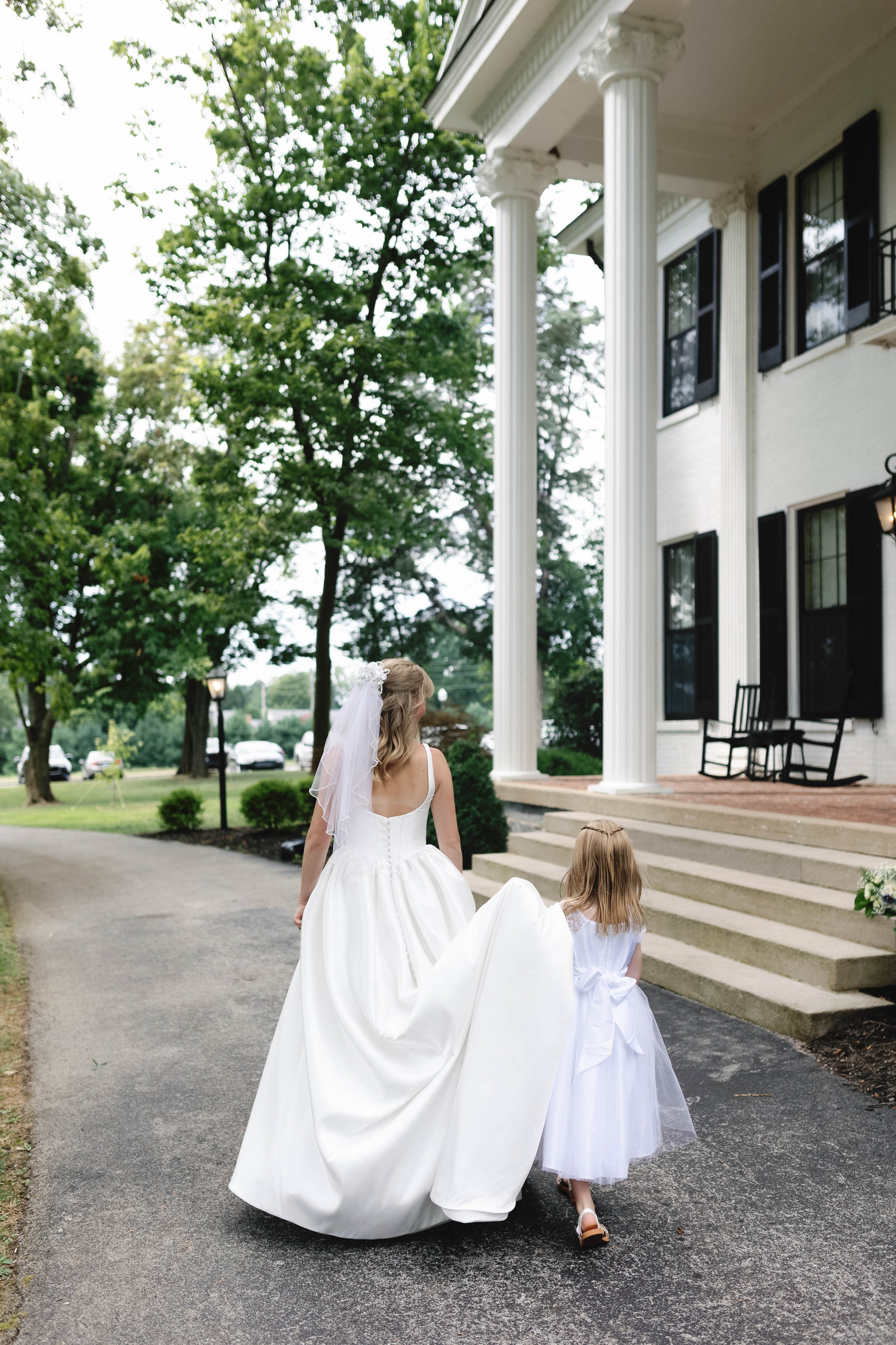 A bride and a young girl walking up the steps of a white house with large columns, surrounded by green trees, on a cloudy day. ashford acres inn wedding cynthiana kentucky