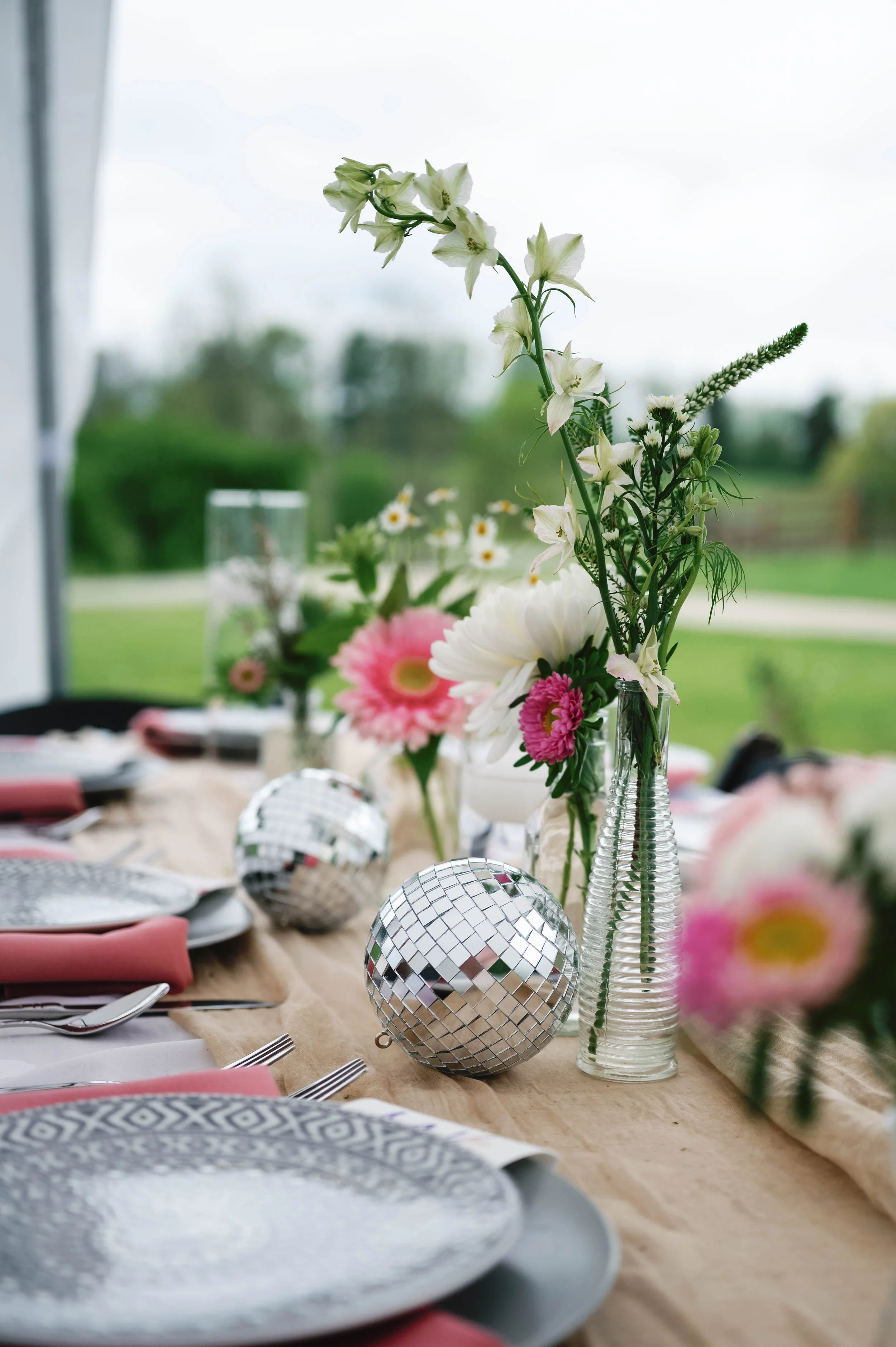 Close-up of a table decorated with pink and white flowers in glass vases, silver disco ball ornaments, plates, napkins, and cutlery, with an outdoor green landscape visible through a window in the background spring wedding in kentucky queenslake bnb
