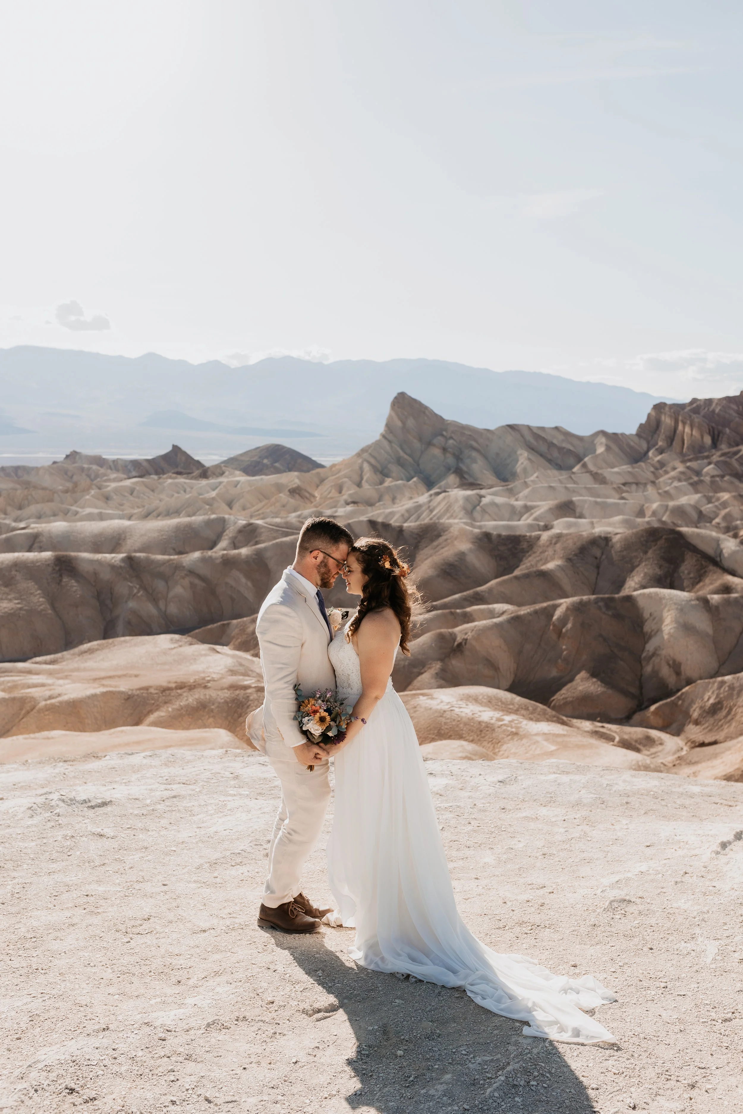 A bride and groom embrace and touch foreheads in a desert landscape with rocky hills and mountains in the background. death valley national park elopement