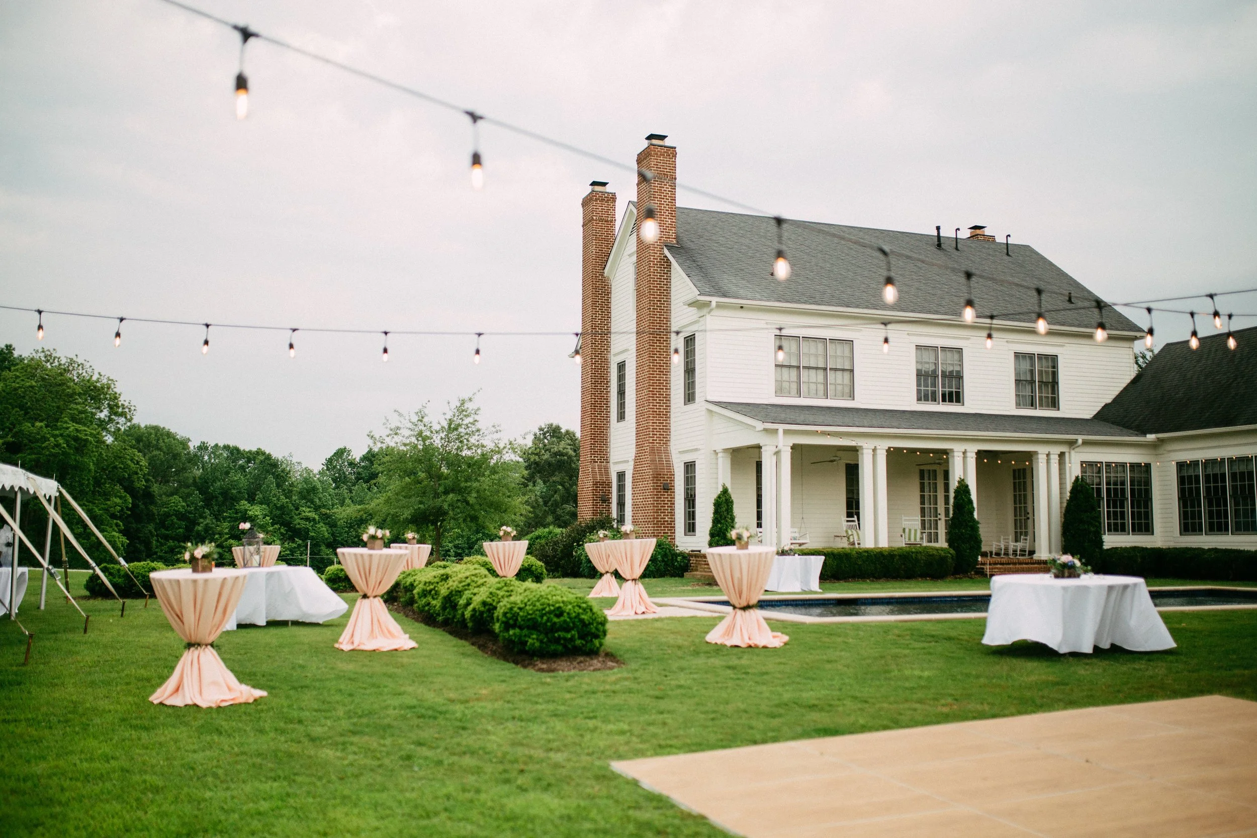 An outdoor yard party setup at a large white house, with string lights overhead, round tables with pink tablecloths and floral centerpieces, and a white tent on the left side Mississippi historic wedding venue