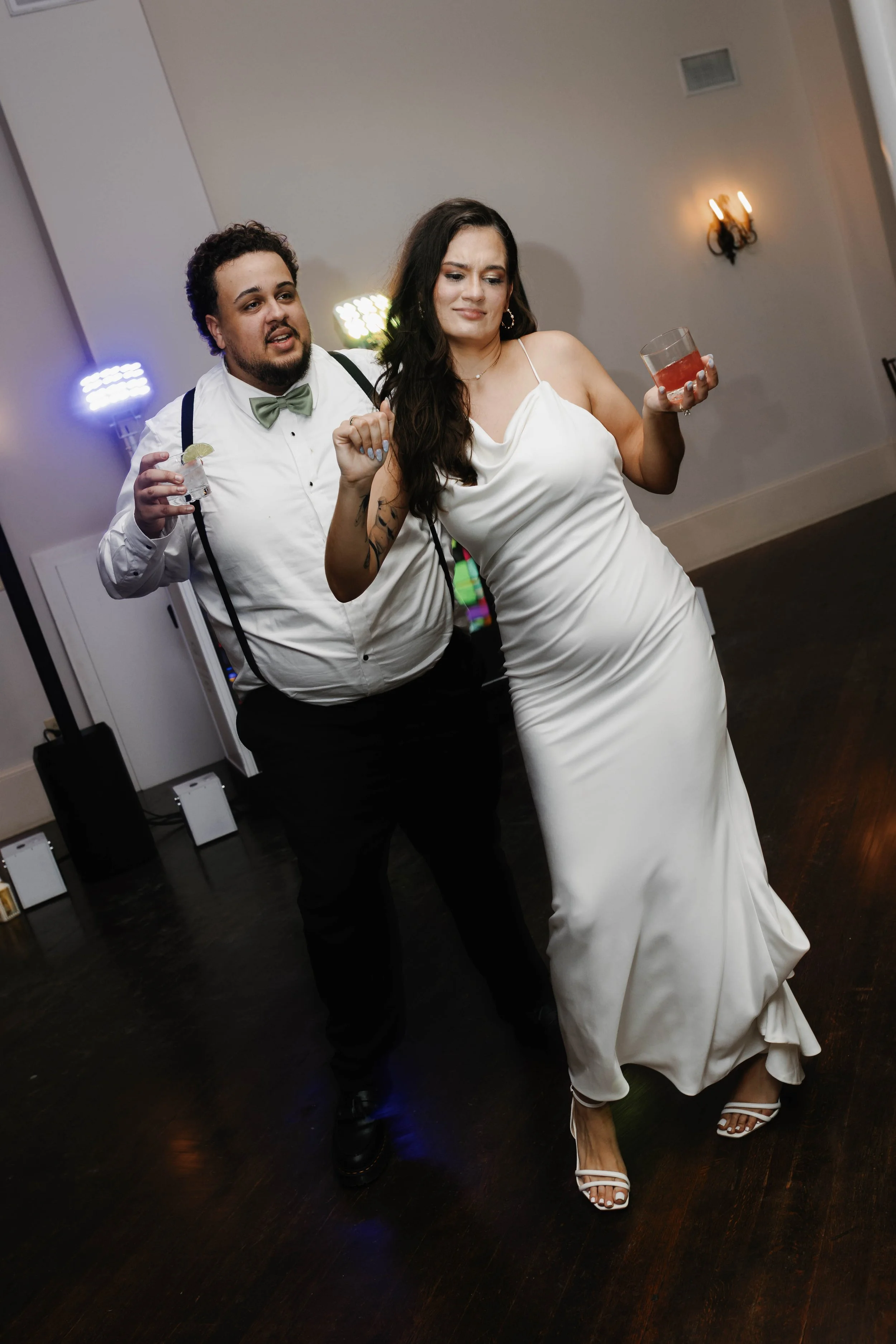 A wedding reception scene with a man in a white shirt, bow tie, and suspenders dancing with a woman in a white wedding gown, both holding drinks, on a dark wood floor with colorful lights and a wall sconce in the background. winchester kentucky