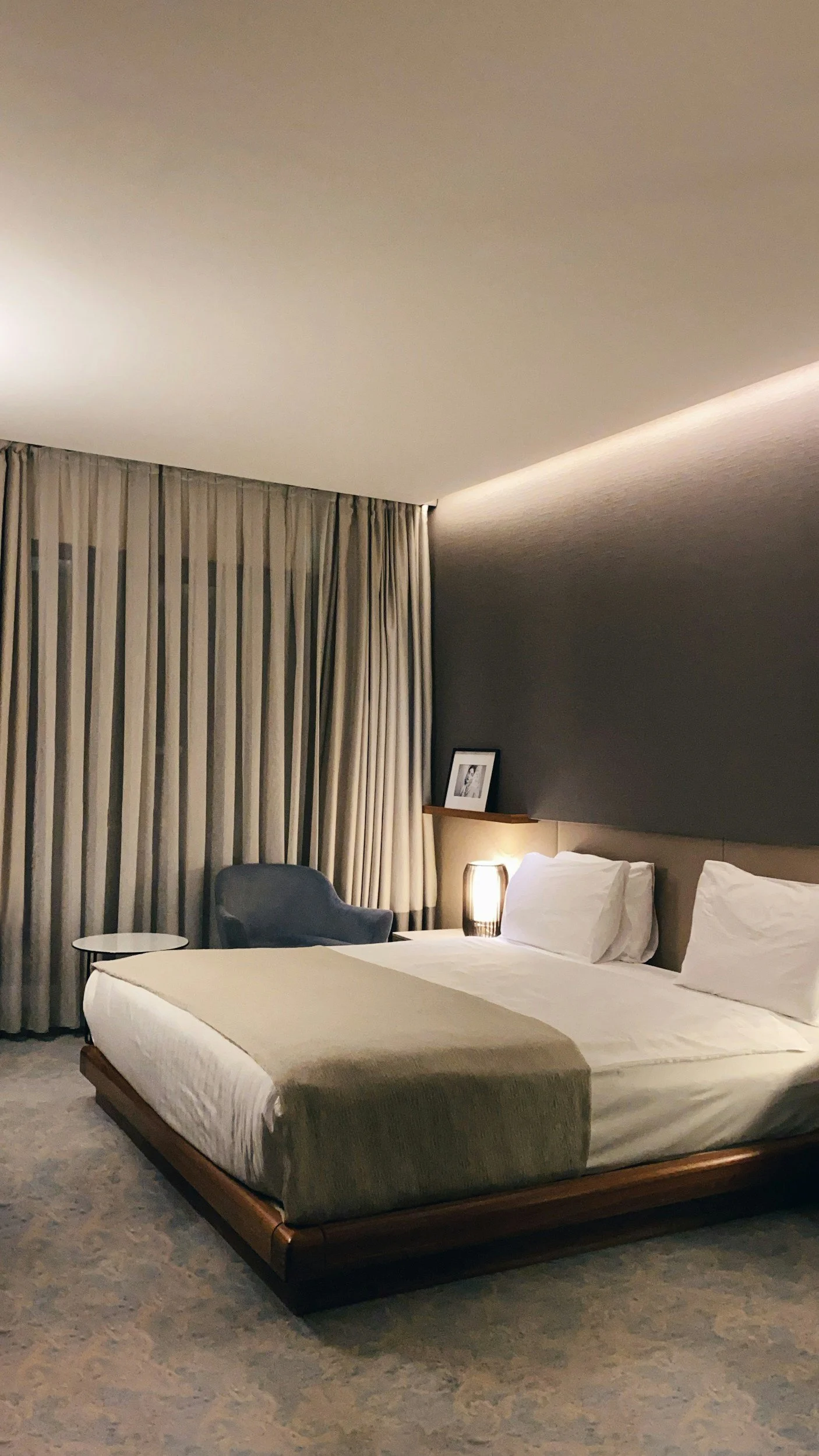 A modern hotel room with a large bed, bedside lamp, armchair, side table, and floor-length curtains.