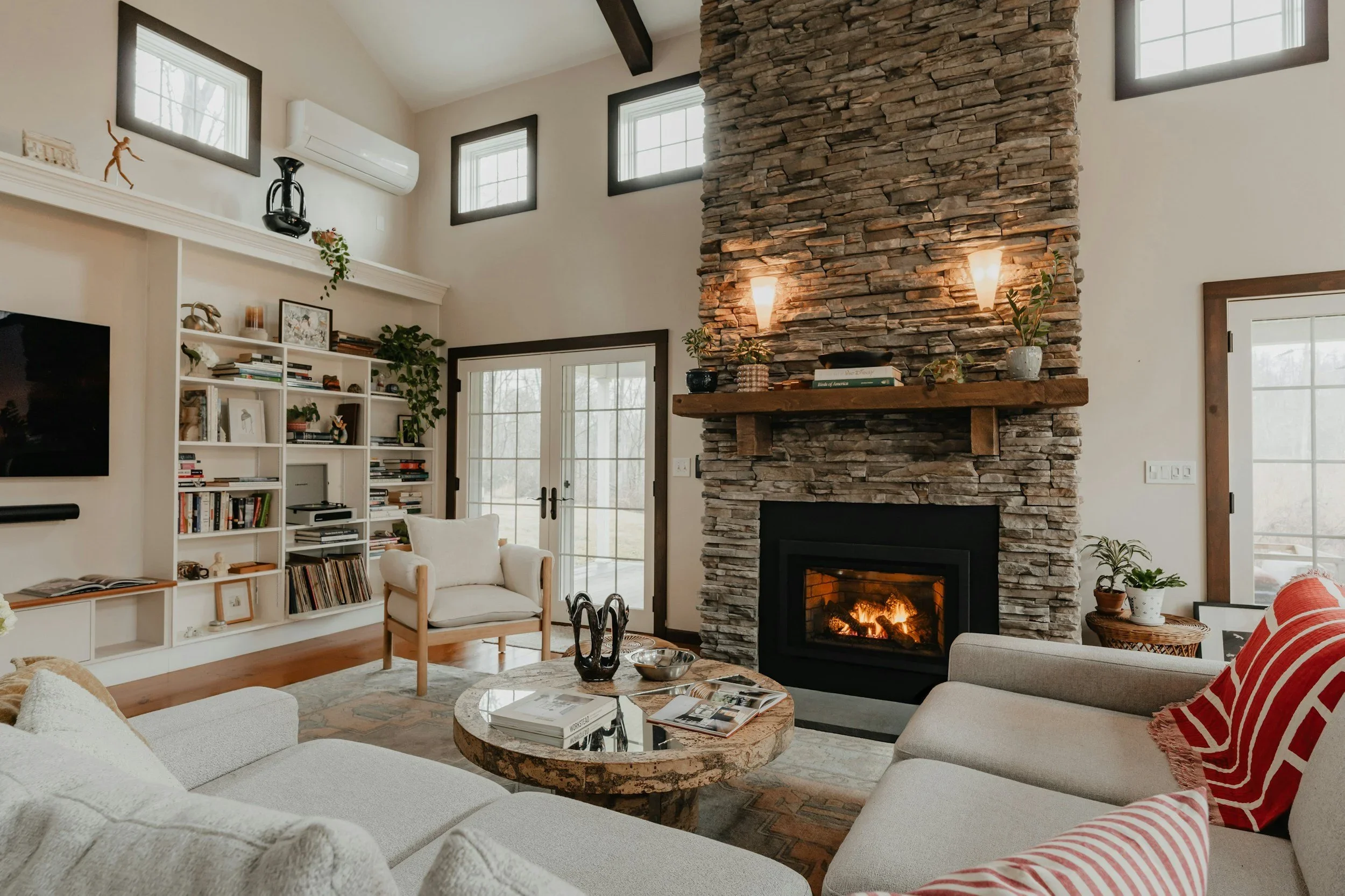 Living room with a stone fireplace, beige sofas, a round coffee table, and large windows.