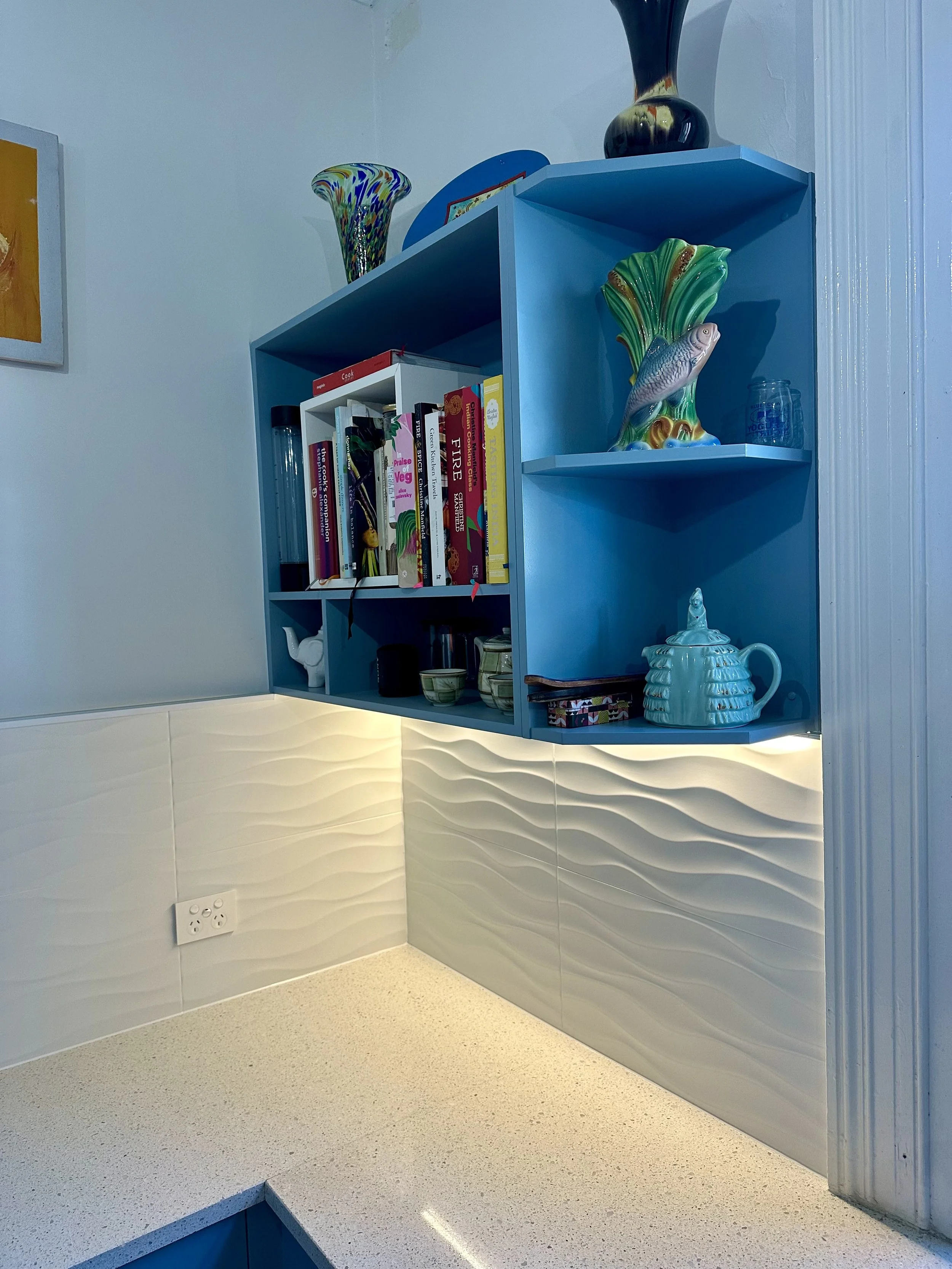 Blue kitchen shelf with books and decorative items, under cabinet lighting, beige countertop, electrical outlet on wall.