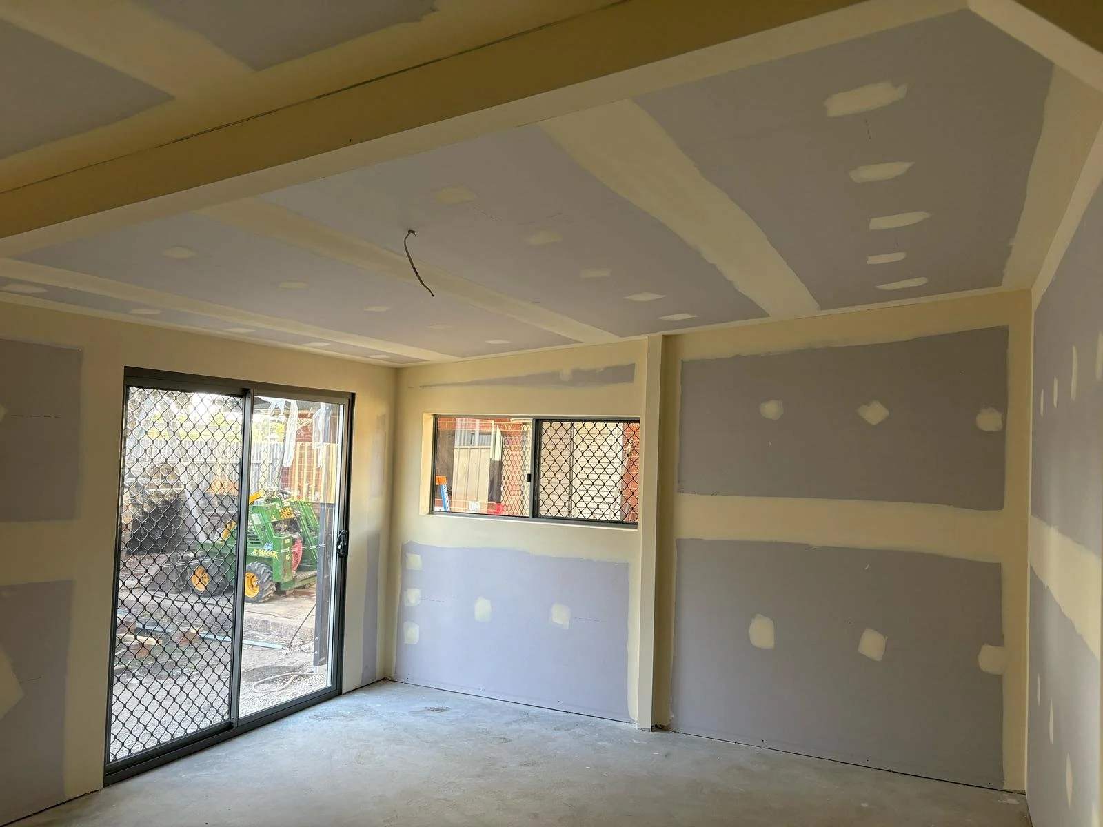 Interior of a room under construction with drywall and unpainted walls, featuring large sliding glass doors and sectioned windows, with visible construction materials outside.