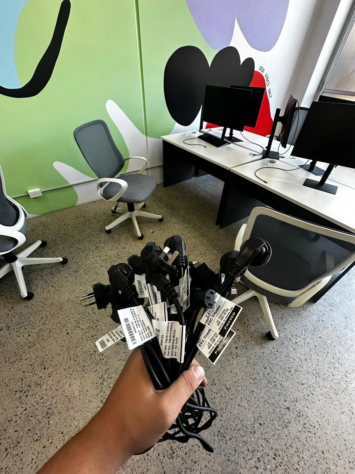 A person is holding multiple black power cords with labels in an office space with two empty office chairs, two computer monitors on desks, and a colorful mural on the wall.