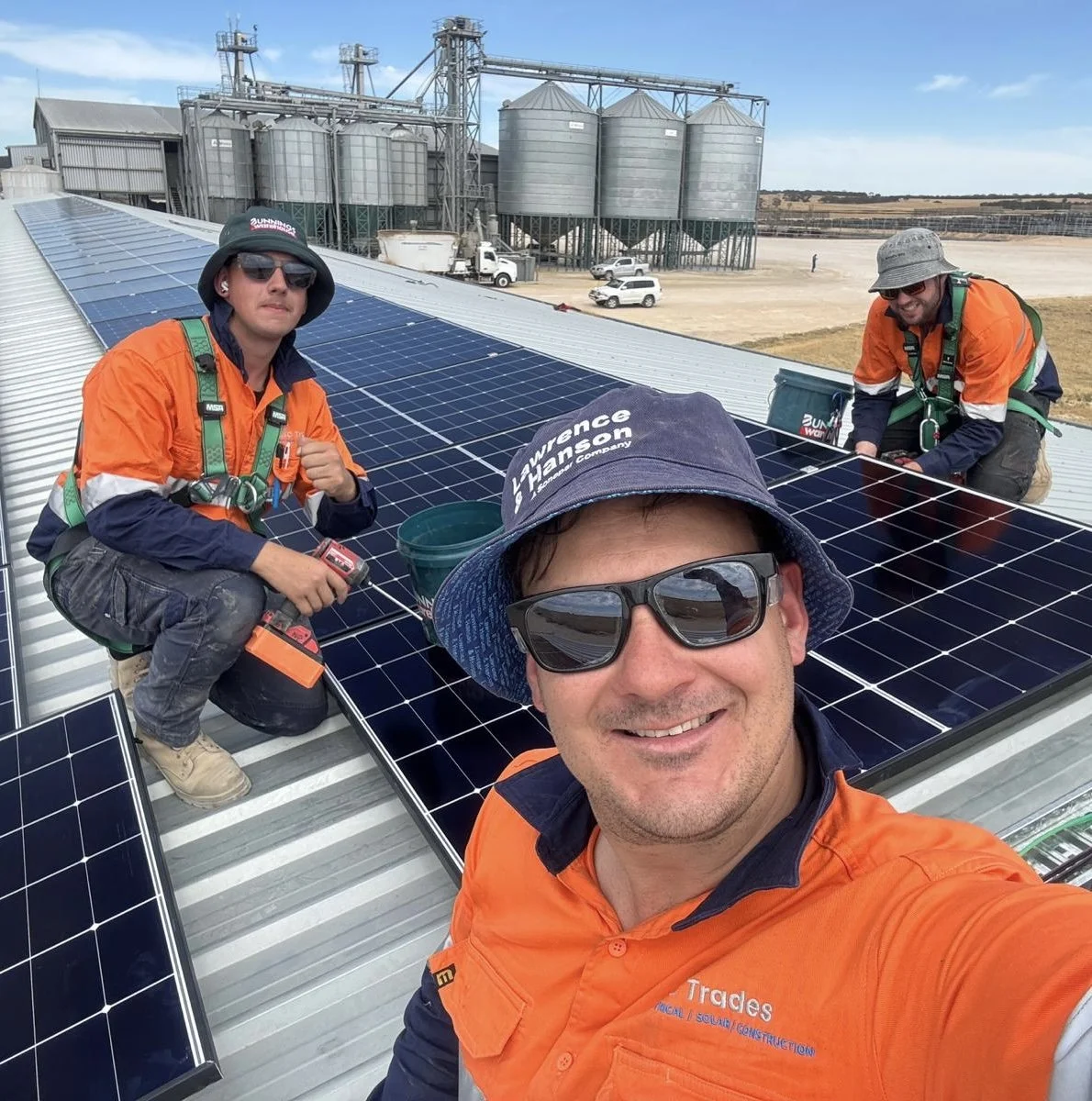 Three workers installing solar panels on a metal roof, with industrial silos and a clear blue sky in the background.