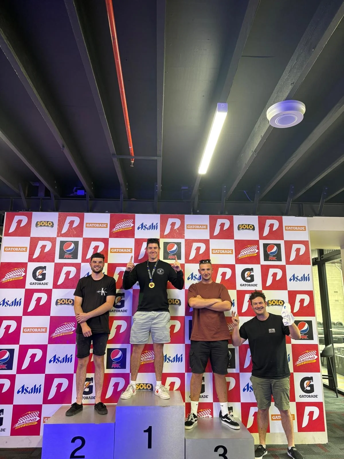 Four men standing on a winners' podium at a racing event, with a backdrop displaying various sponsor logos including Gatorade, Asahi, Solo, and Pea. The man in the center is on the first-place platform, holding up a thumbs-up and wearing a medal. The others are on the second and third-place platforms, with one man making a peace sign and another smiling while holding a paper towel.