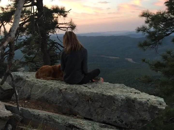 A woman and a dog sitting on a rocky ledge overlooking a mountain landscape at sunset, with trees framing the scene.