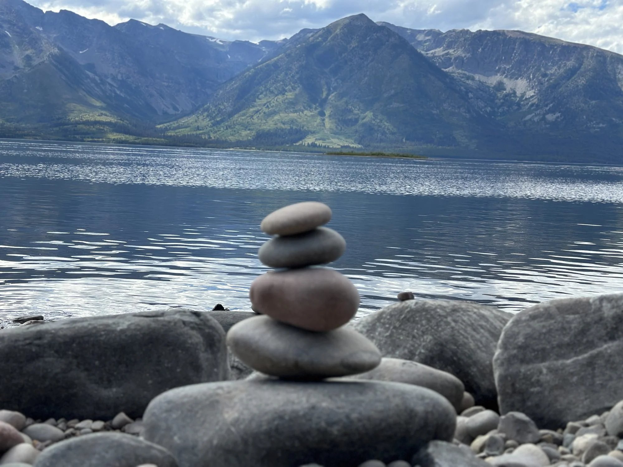 Stacked smooth stones on a rocky lakeshore with a mountain range in the background under a partly cloudy sky.