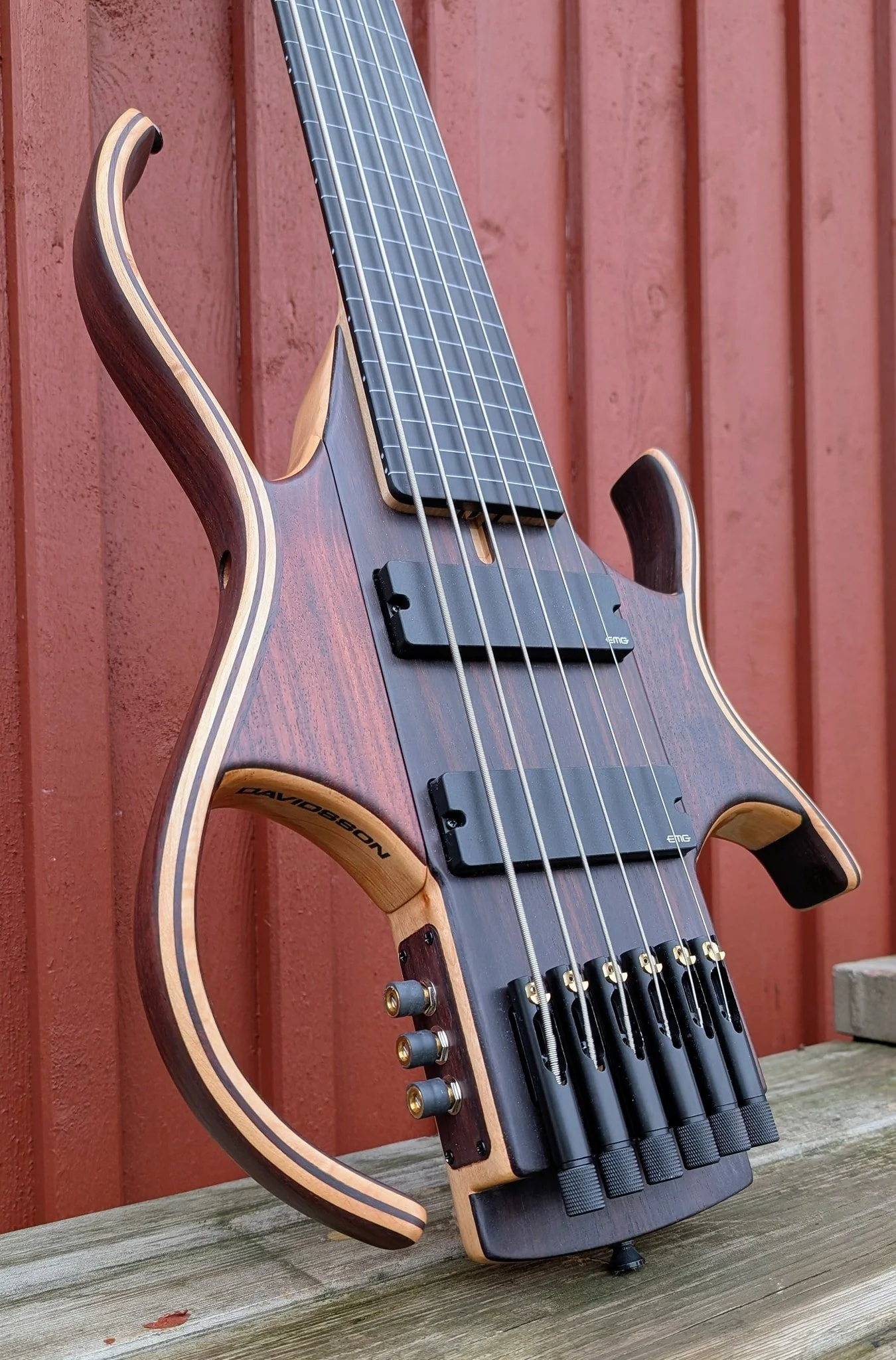 Elektrisk basgitarr med träfinish, utomhus mot en rödfärgad trävägg.