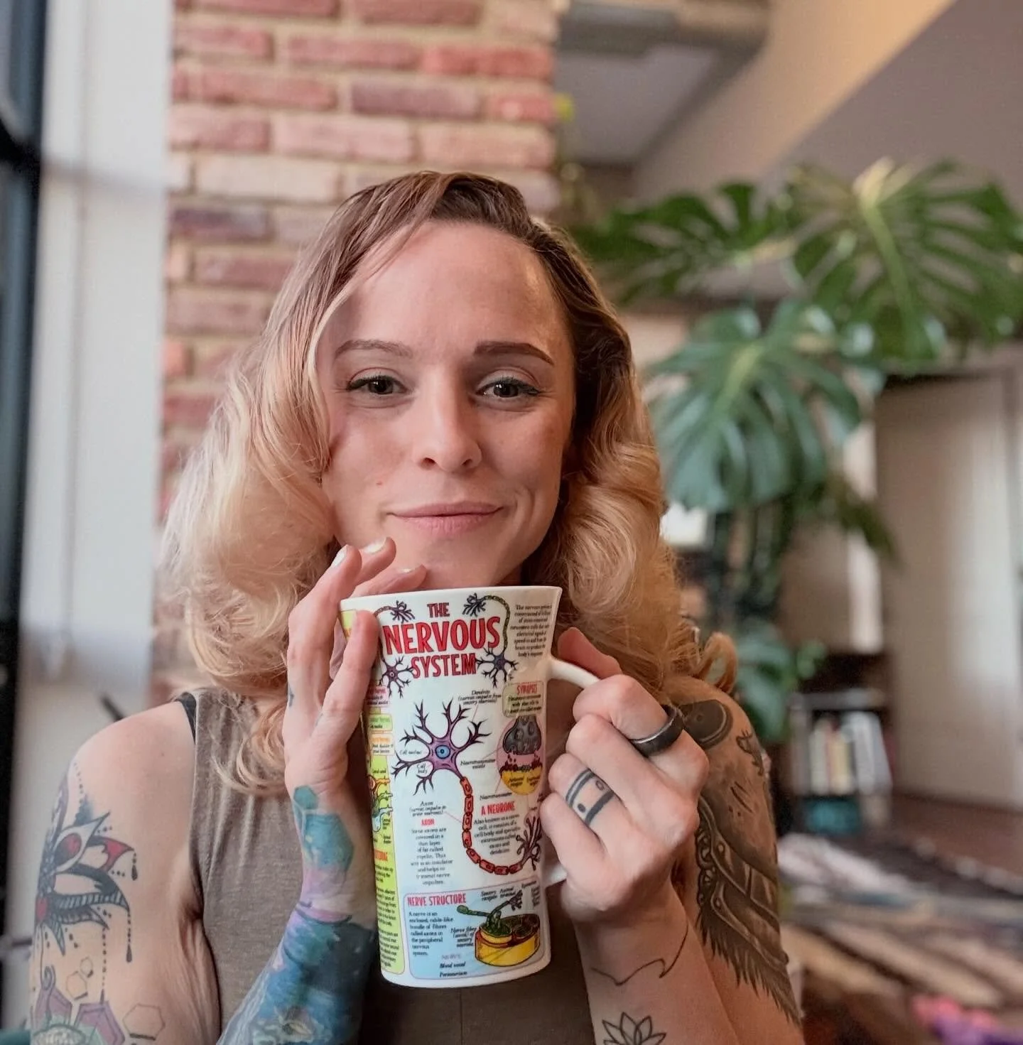 A woman with tattoos on her arms holding a coffee mug that reads 'The Nervous System' with an anatomical diagram of the nervous system printed on it. She is smiling slightly and has wavy blonde hair. There is a brick wall and a large potted plant in the background.