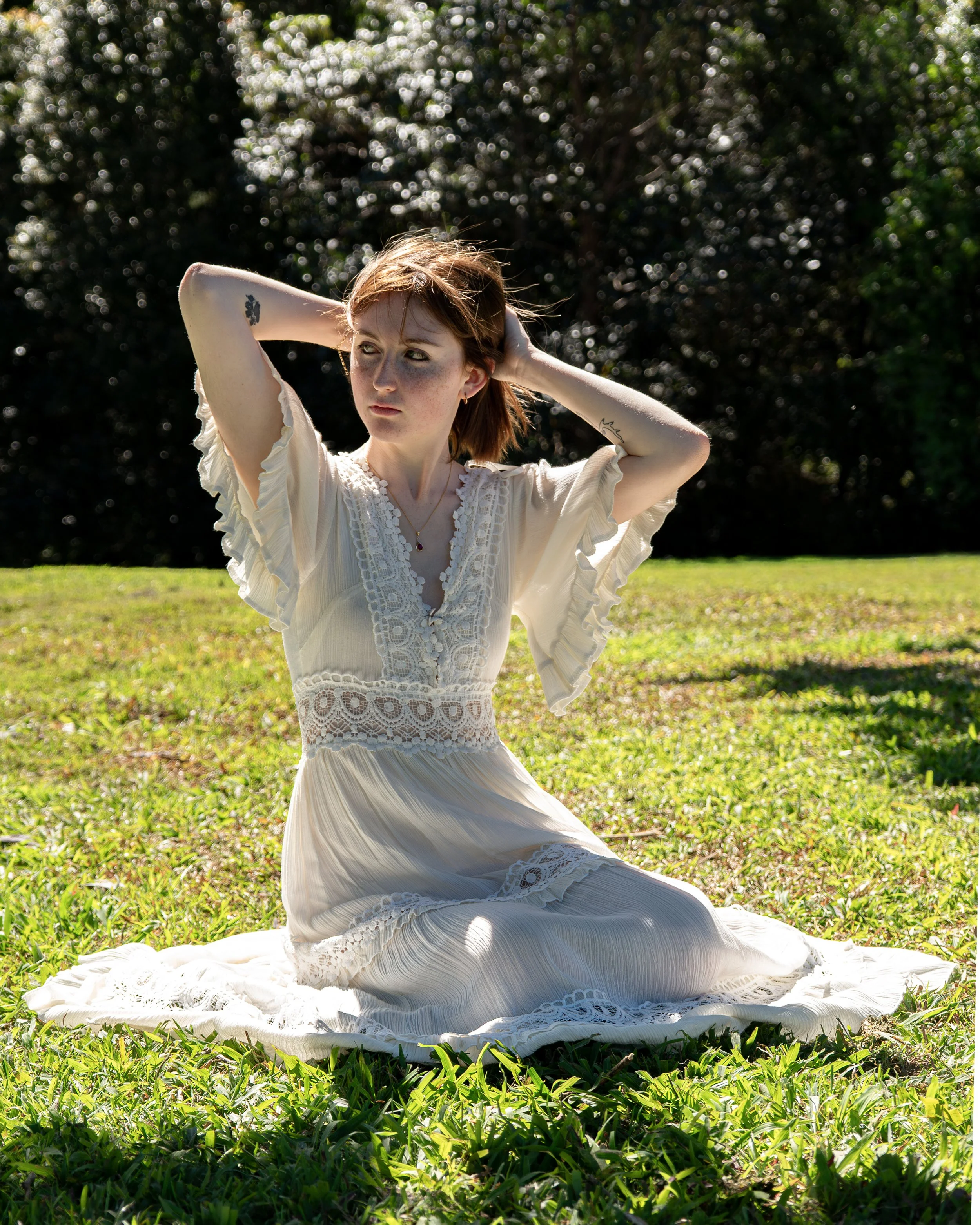 A woman in a white dress sitting on grass with her hands behind her head, in a natural outdoor setting with trees in the background.