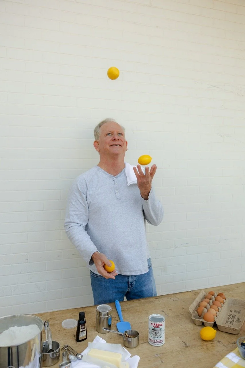 An elderly man in a gray shirt and jeans juggling three yellow lemons in a kitchen or baking area, with eggs, utensils, and ingredients on the table.