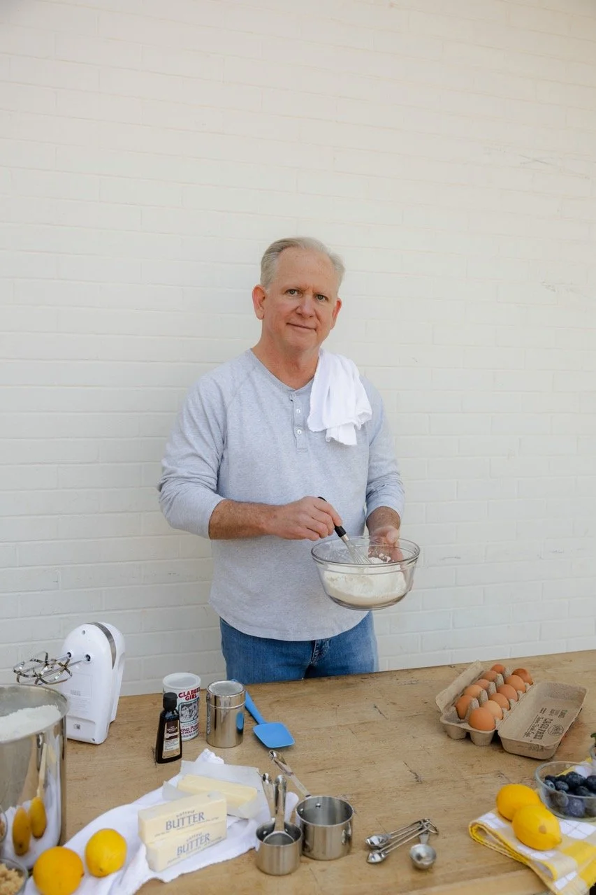 A man with gray hair, wearing a light gray long-sleeve shirt and blue jeans, stands behind a wooden table with baking ingredients and utensils. He is mixing batter in a glass bowl, with a white towel hanging around his neck. On the table, there are eggs, butter, blueberries, lemons, a hand mixer, measuring cups, and baking tools, against a white brick wall background.