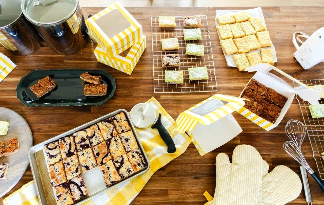 Assorted baked bars and cookies on cooling racks, plates, and boxes on a wooden table, with baking tools and oven mitts nearby.