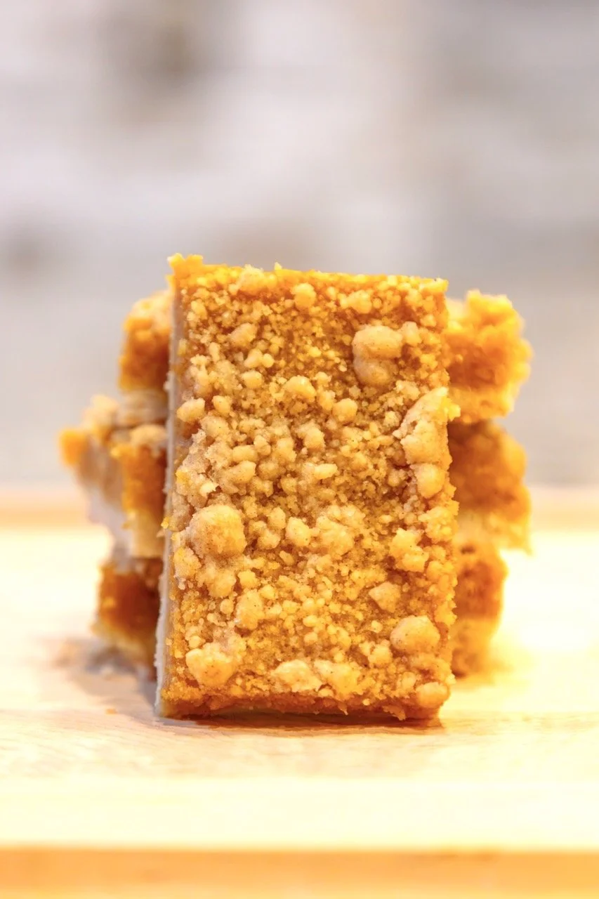 Close-up of two stacked crumbly, golden-brown snack bars with a textured surface on a light wooden surface.