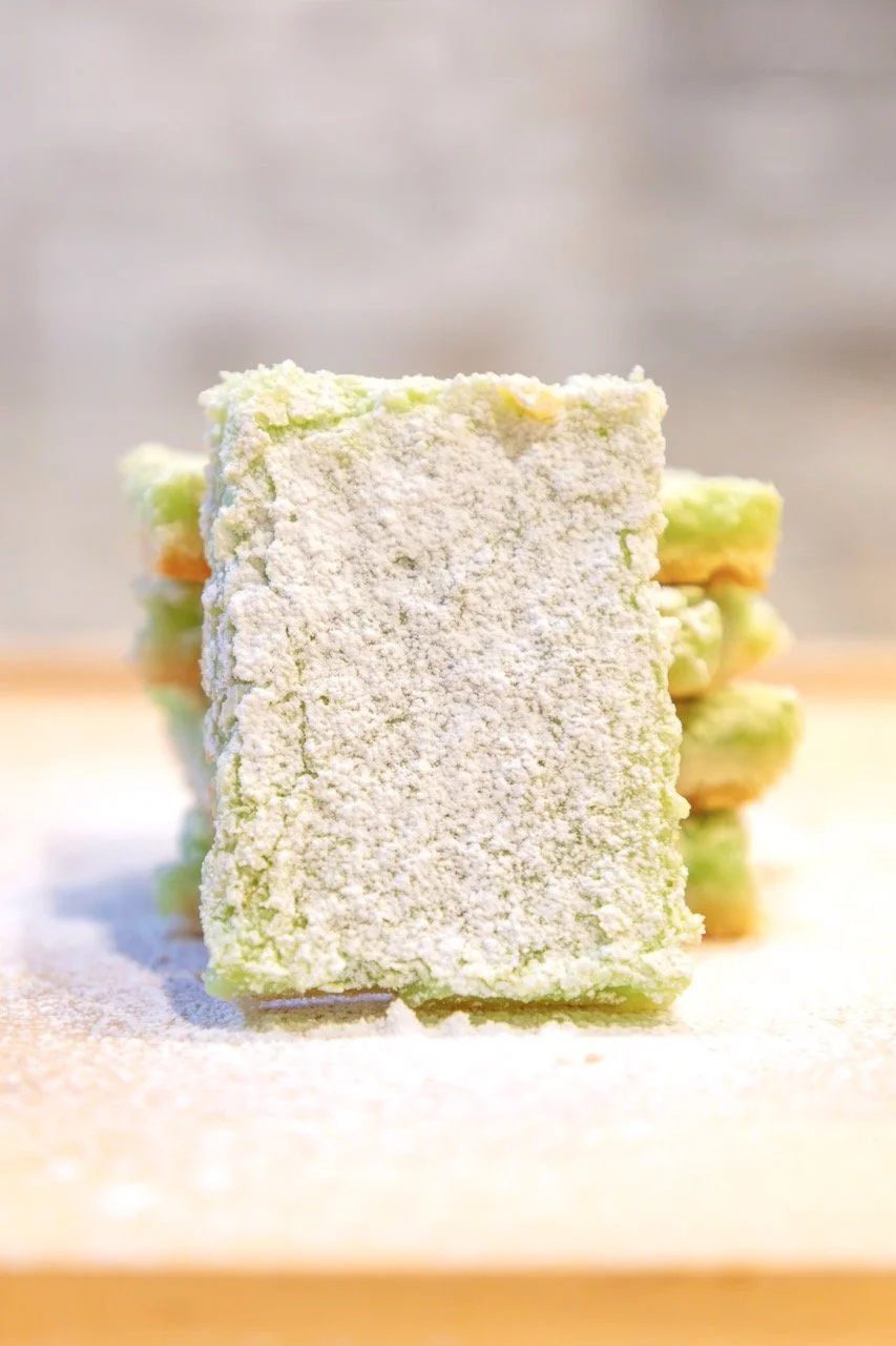Stack of layered matcha-flavored dessert bars dusted with powdered sugar on a wooden surface.