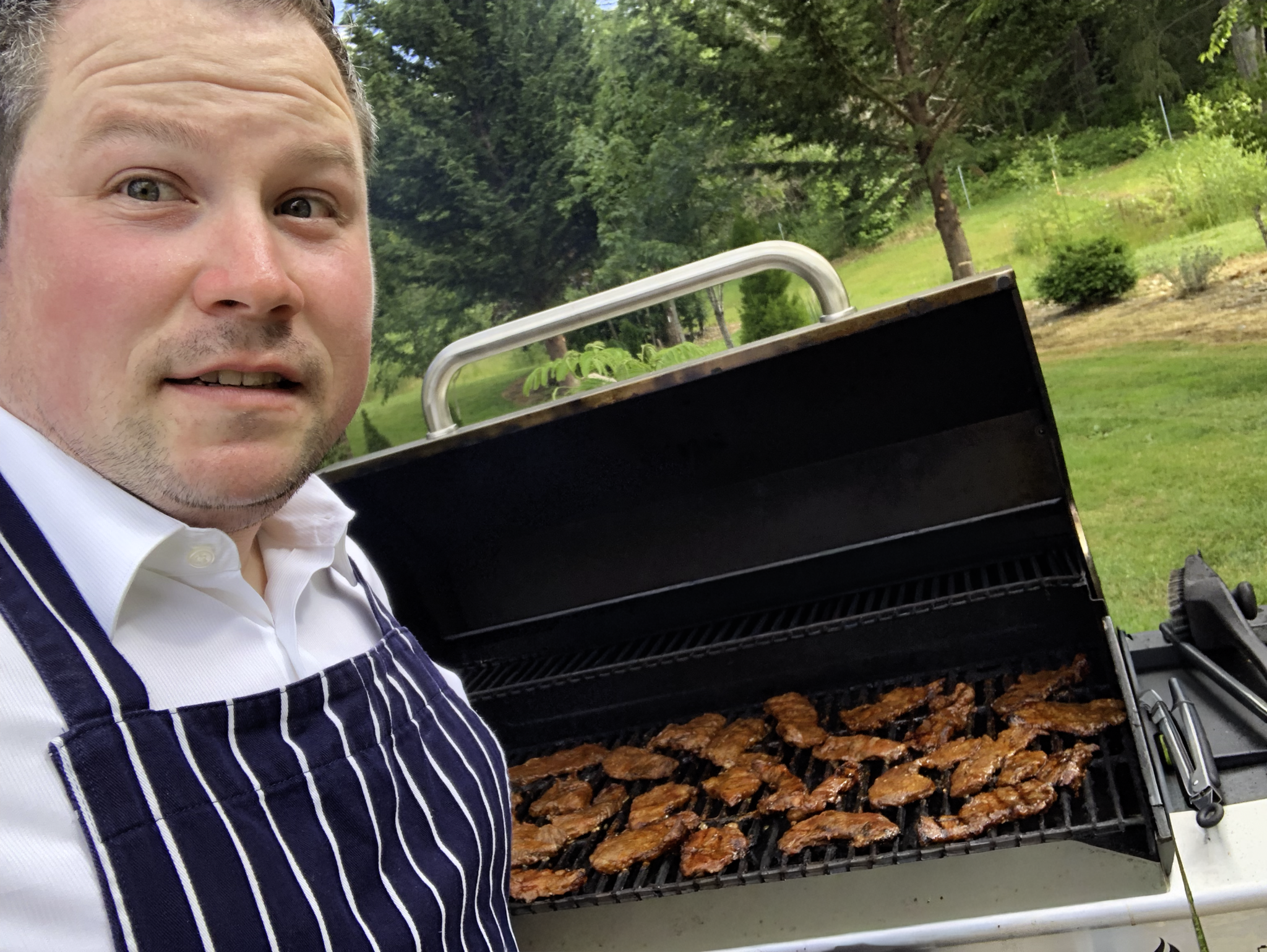 A man in a white shirt and striped apron takes a selfie beside an outdoor grill filled with cooked meat, with trees and grass in the background.