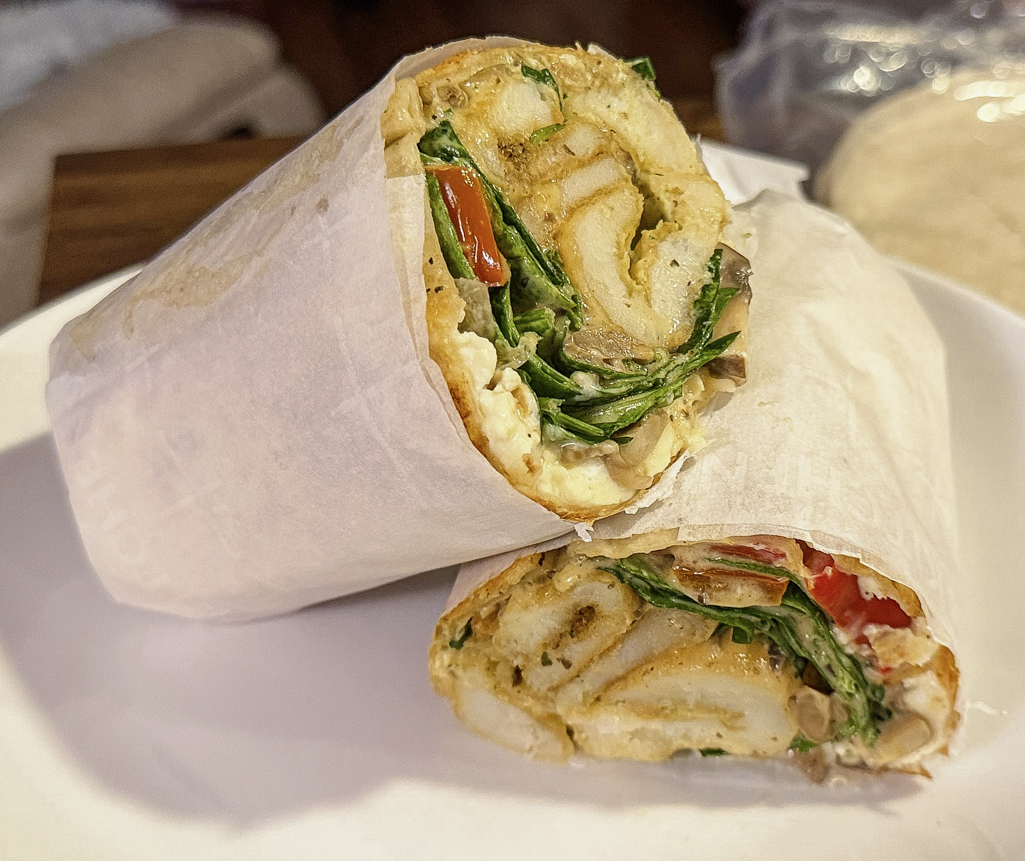 Close-up of a veggie burrito sliced in half, wrapped in white paper, showing lettuce, tomato, onion, eggs, sidewinder pototaes and grilled vegetables inside.