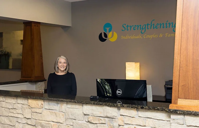 A woman with gray hair and a black sweater standing behind a stone reception desk in an office lobby, with a large computer monitor in front of her and a lit lamp behind it, and a logo on the wall that reads 'Strengthening Individuals, Couples & Families'.