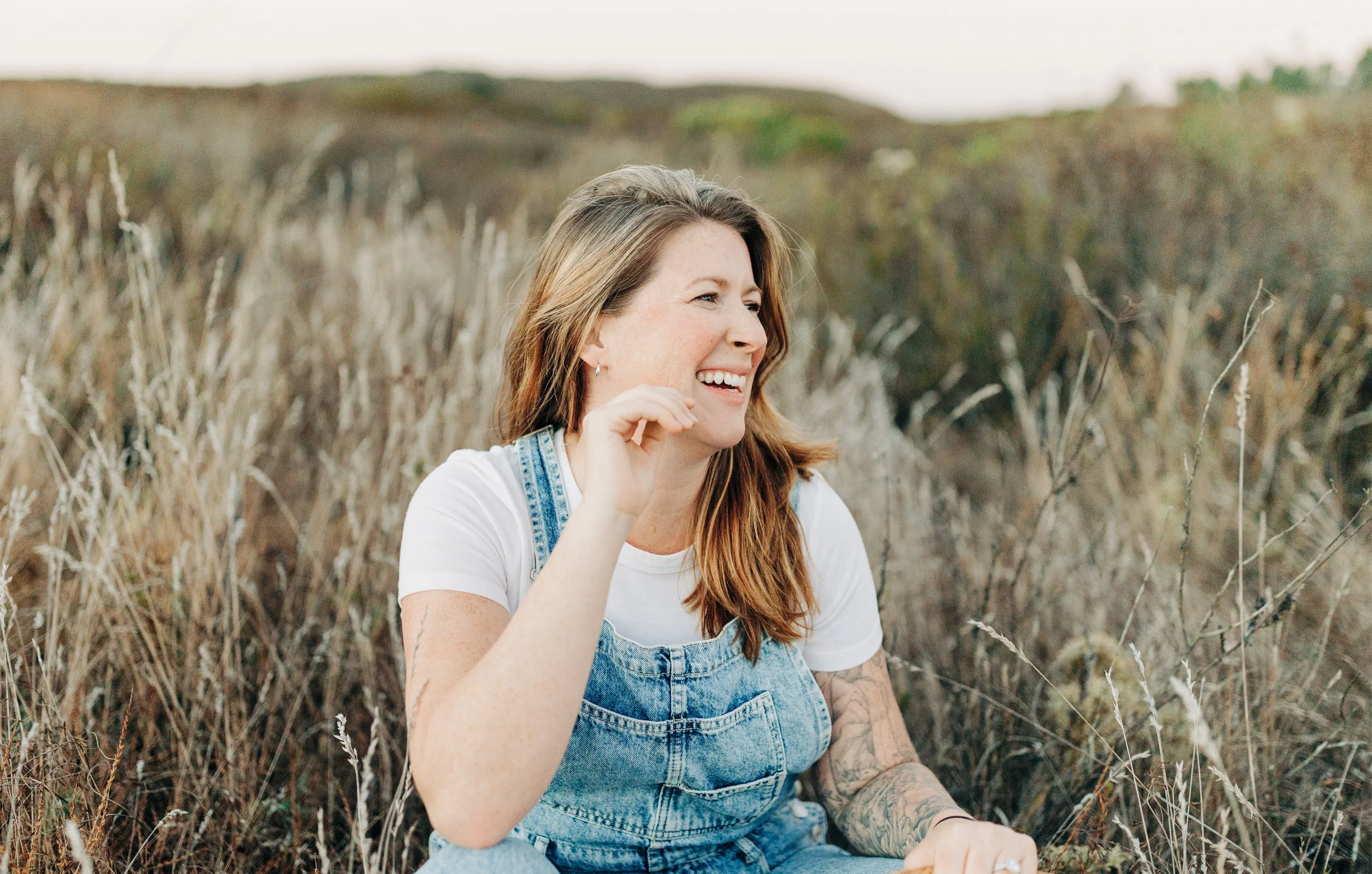 A woman smiling and sitting in a field of tall, dry grass, wearing a white t-shirt and denim overalls.